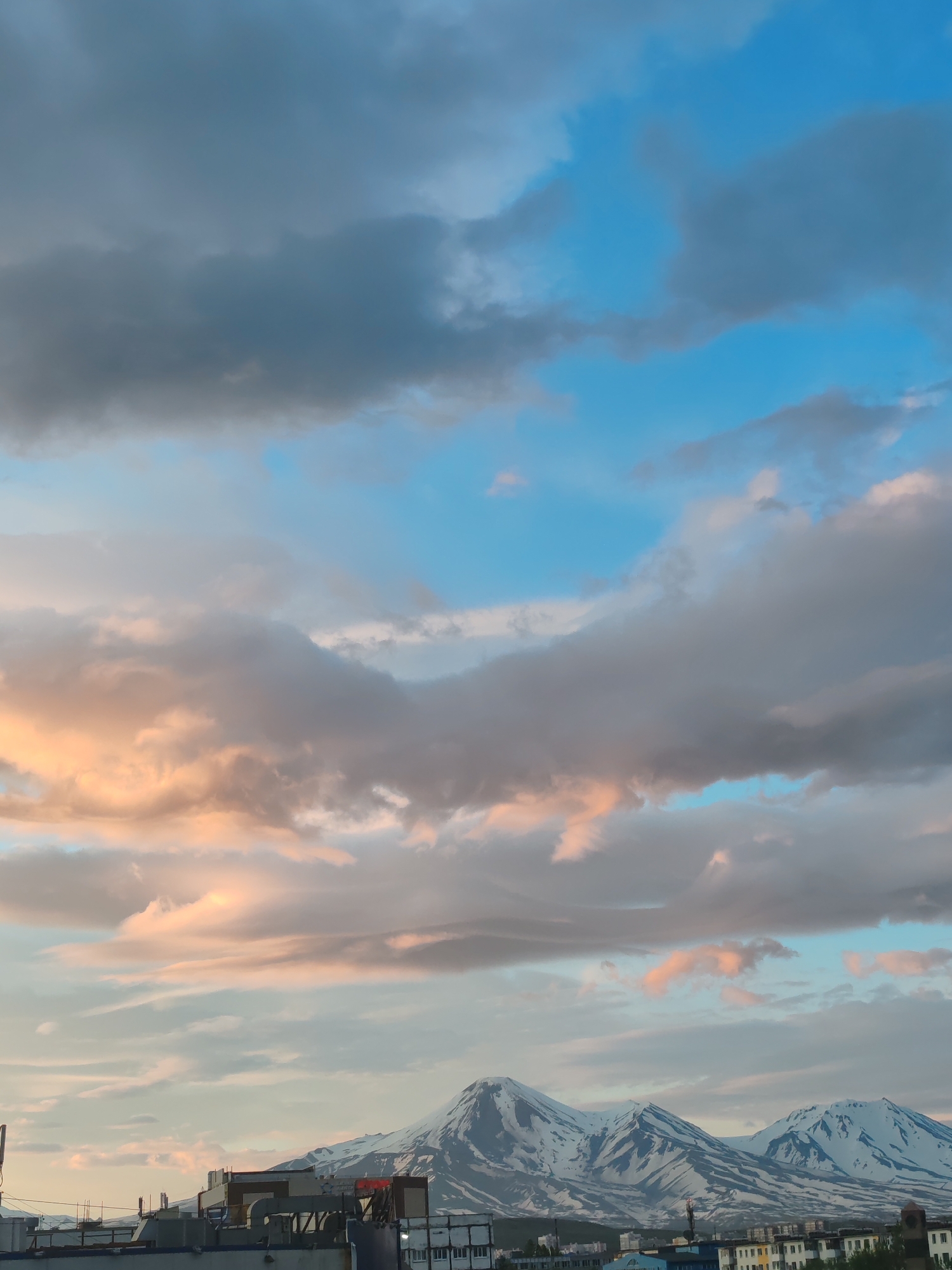 The image depicts a broad view of the city of Petropavlovsk-Kamchatsky, Russia, with a prominent mountain range in the background. The sky is filled with dramatic clouds, some tinged with the warm hues of sunset or sunrise, contrasting with areas of clear blue. The foreground shows a cityscape with buildings, including industrial-looking structures with pipes and roofs, suggesting a developed urban area. Further in the background, the majestic snow-capped mountains rise above the city, their slopes streaked with dark rock and snow. The overall impression is one of a city nestled beneath imposing natural landscapes, with the dynamic sky adding to the scene's atmosphere.