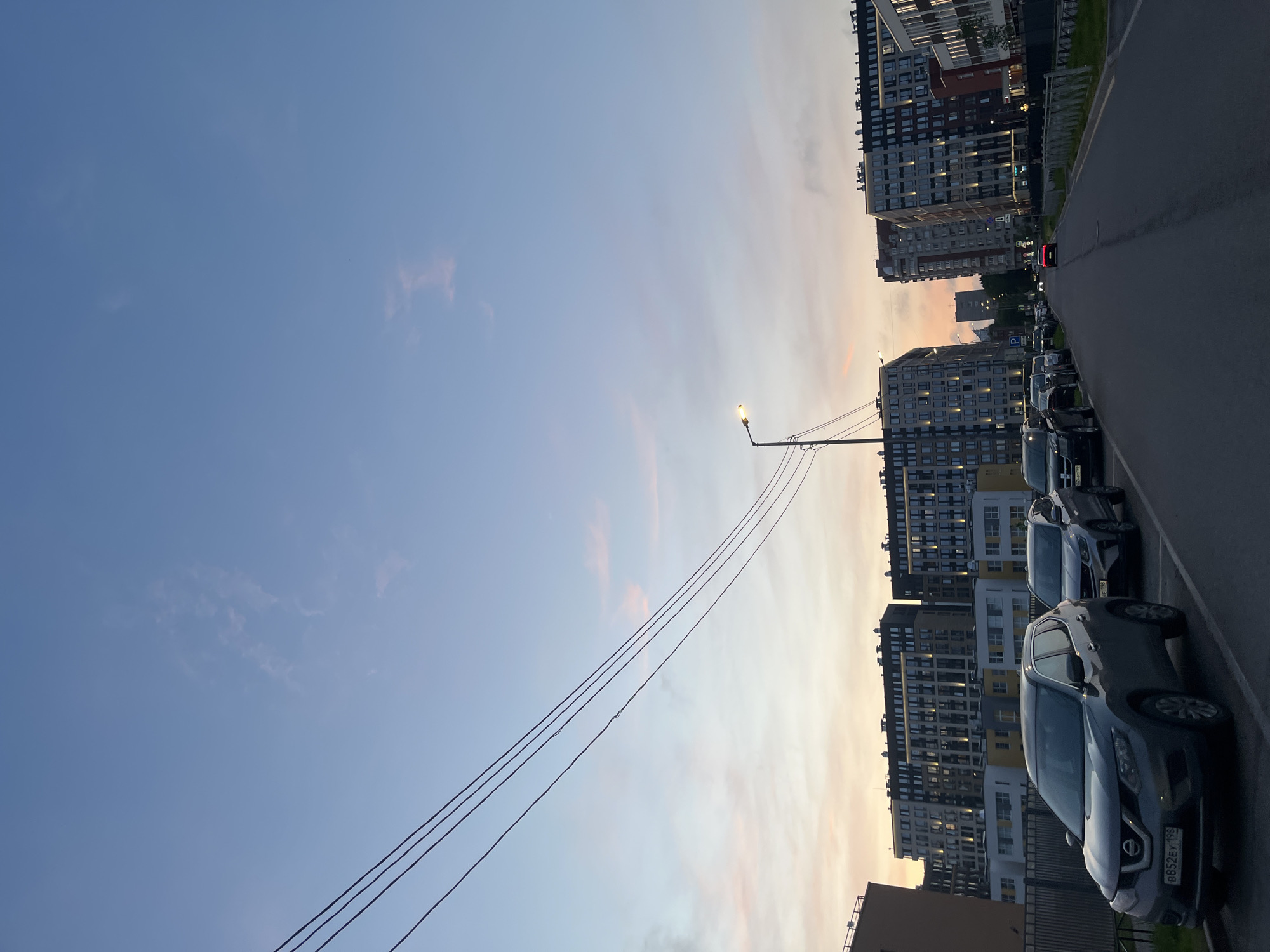 The image depicts a street scene in Saint Petersburg, Russia, at dusk. Buildings of various heights line the street, with many windows illuminated. Cars are parked along the right side of the road. A street lamp is visible with power lines extending across the frame. The sky shows a gradient of blue transitioning to orange near the horizon. No people are visible in the scene. The overall setting suggests an urban residential area.