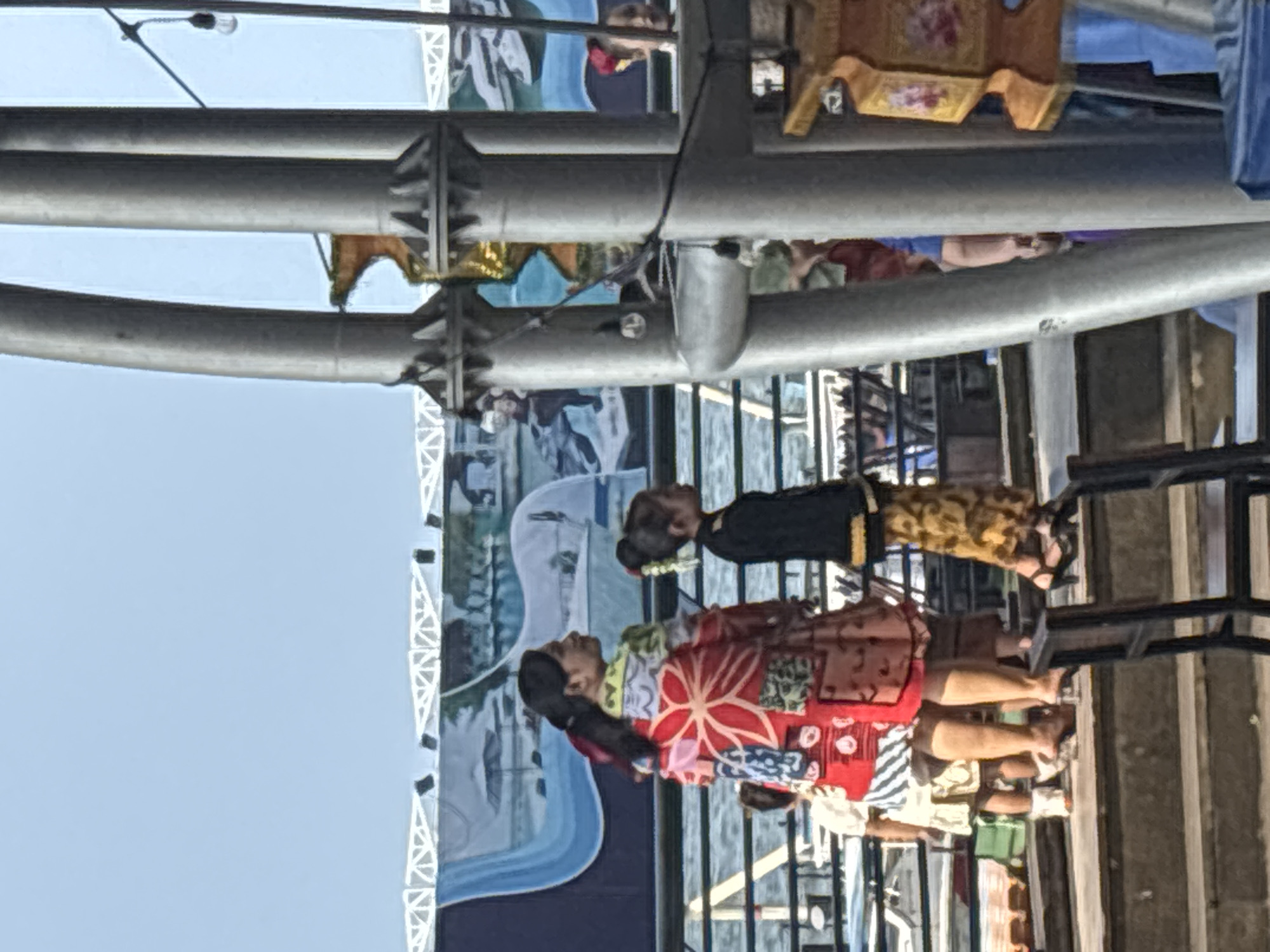 Two women dressed in traditional clothing stand in front of a display screen, possibly at an outdoor event in North Jakarta, Indonesia. One wears a red and white patterned kimono-style garment, while the other wears a black and brown patterned dress. Steel support beams and metal fencing are visible in the foreground, obscuring some of the scene. There are spectators behind them in the middleground. A banner with a blue and white design is behind the figures. The sky is visible in the upper left.