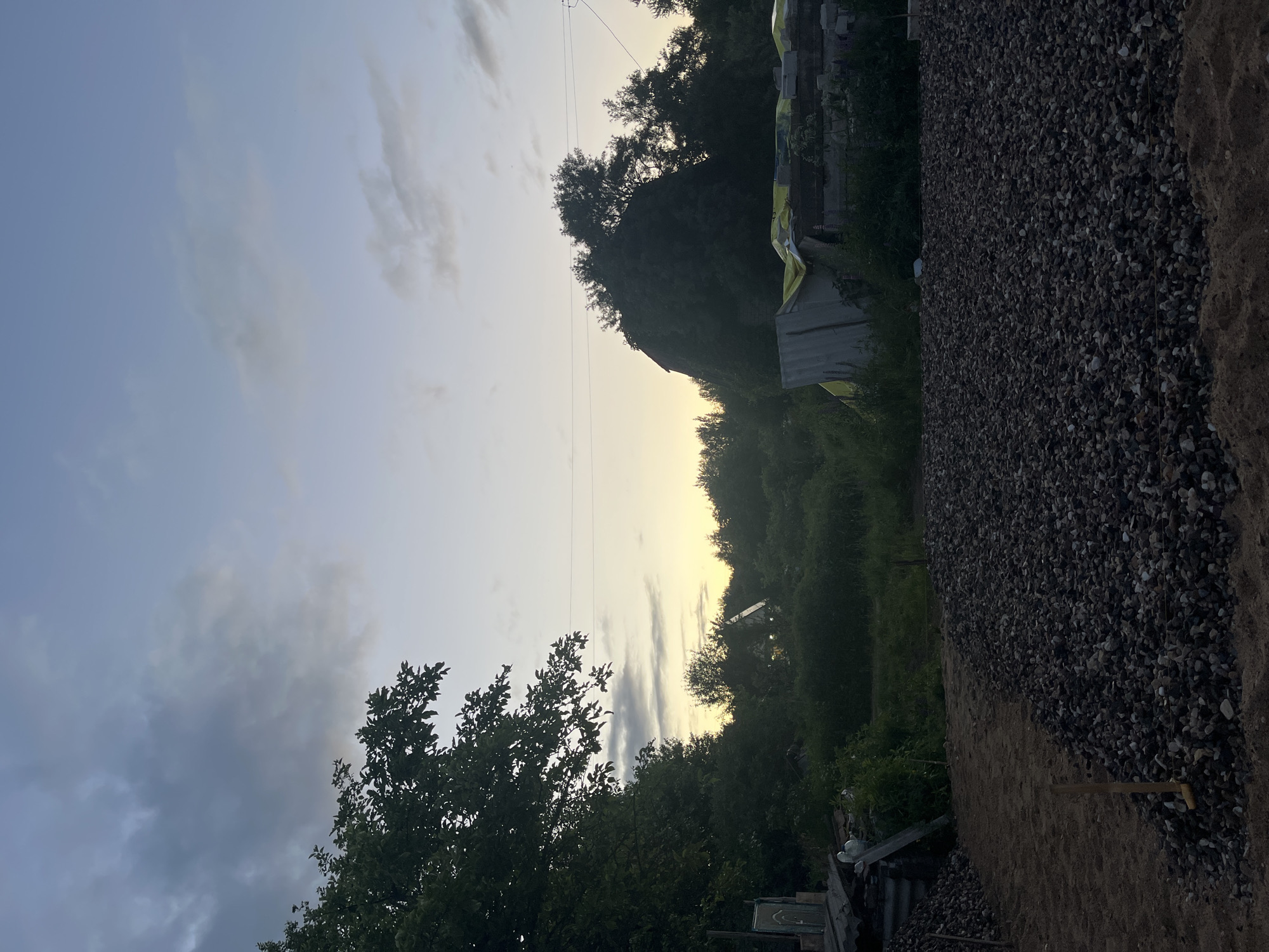 The image depicts an outdoor scene at dusk, possibly in the village of Klyobushnevo, Russia. In the foreground, a large pile of dark grey gravel is visible, with a section of lighter sand next to it. A wooden stake is partially embedded in the gravel. Behind the gravel, there is a dense area of green foliage and trees. In the mid-ground, a dilapidated structure with a yellow tarp is visible, and further back, a small, illuminated house with a steeply pitched roof can be seen amongst the trees. The sky above is a gradient of pale blue to light yellow, with scattered dark and light clouds. Power lines are stretched across the upper part of the image. The overall impression is one of rural or semi-developed land at the end of the day.