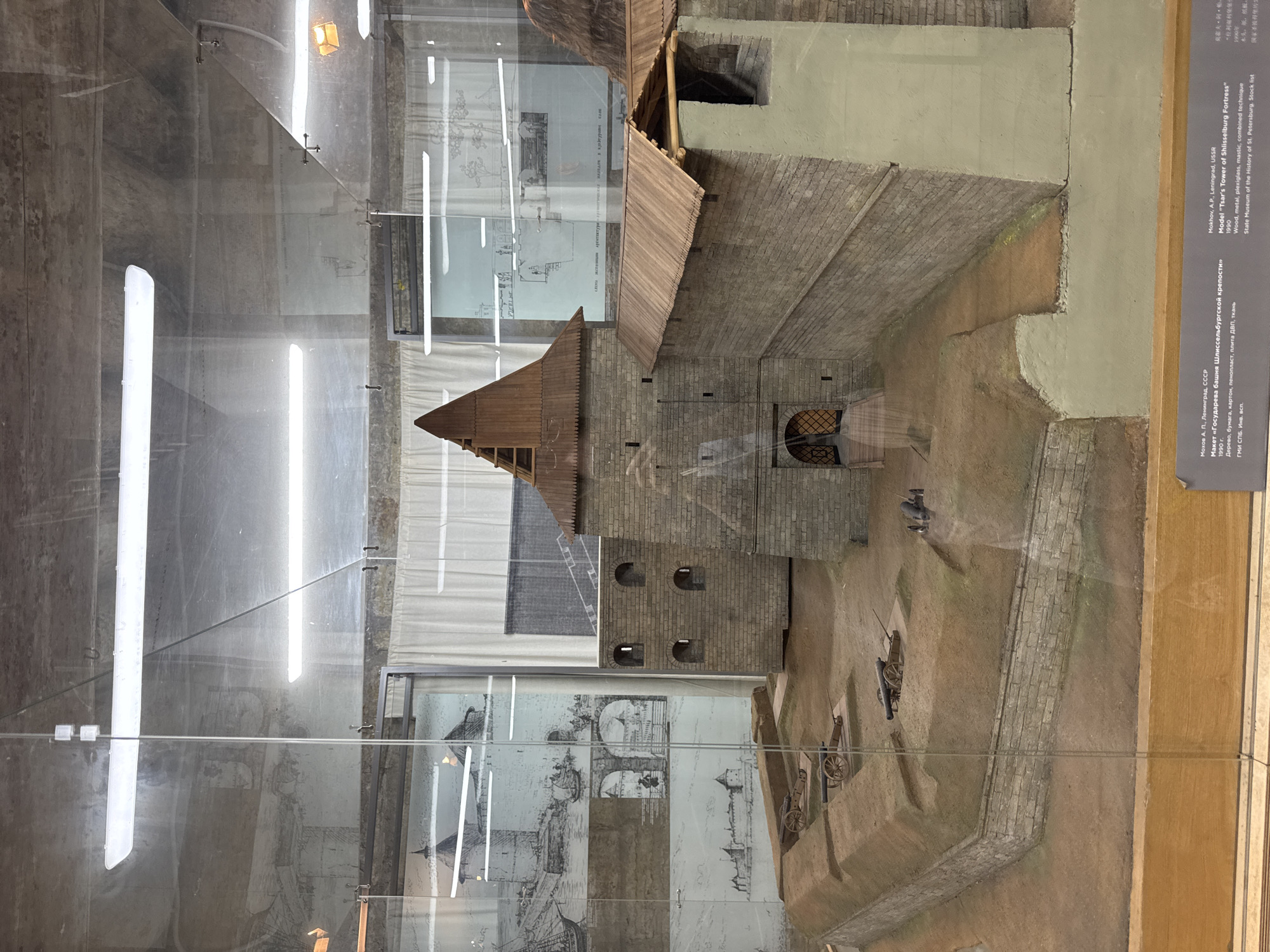 This media file displays a diorama or model of a historical fortress. The model depicts a stone structure with a prominent wooden-roofed tower. The fortress appears to be situated on a gentle slope with a moat-like area. Several miniature cannons are positioned around the exterior.
The background of the diorama includes display panels with historical drawings and blueprints of similar structures, hinting at the model's purpose as an educational exhibit. The lighting suggests an indoor setting, possibly a museum, with fluorescent lights visible reflecting on the protective glass casing.
A text label is visible on the right side of the media file, providing information about the model. The text is in English and Russian, identifying the subject as "Model 'Tsar's Tower of Shlisselburg Fortress'". It also notes the material as "Wood, metal, plexiglass, mastic, combined technique", the artist as "Mokhov A.P.", the location as "Leningrad, USSR", and the year of creation as "1990". It further states that the model is part of the "State Museum of the History of St. Petersburg".
The overall impression is that of a detailed historical reconstruction, likely intended for public viewing and educational purposes, showcasing a significant architectural and military site. There are no people or animals visible in the model itself, and the scene is static, representing a historical representation rather than a dynamic event. The time of day and weather are not discernible as it's a model in an indoor exhibition space.