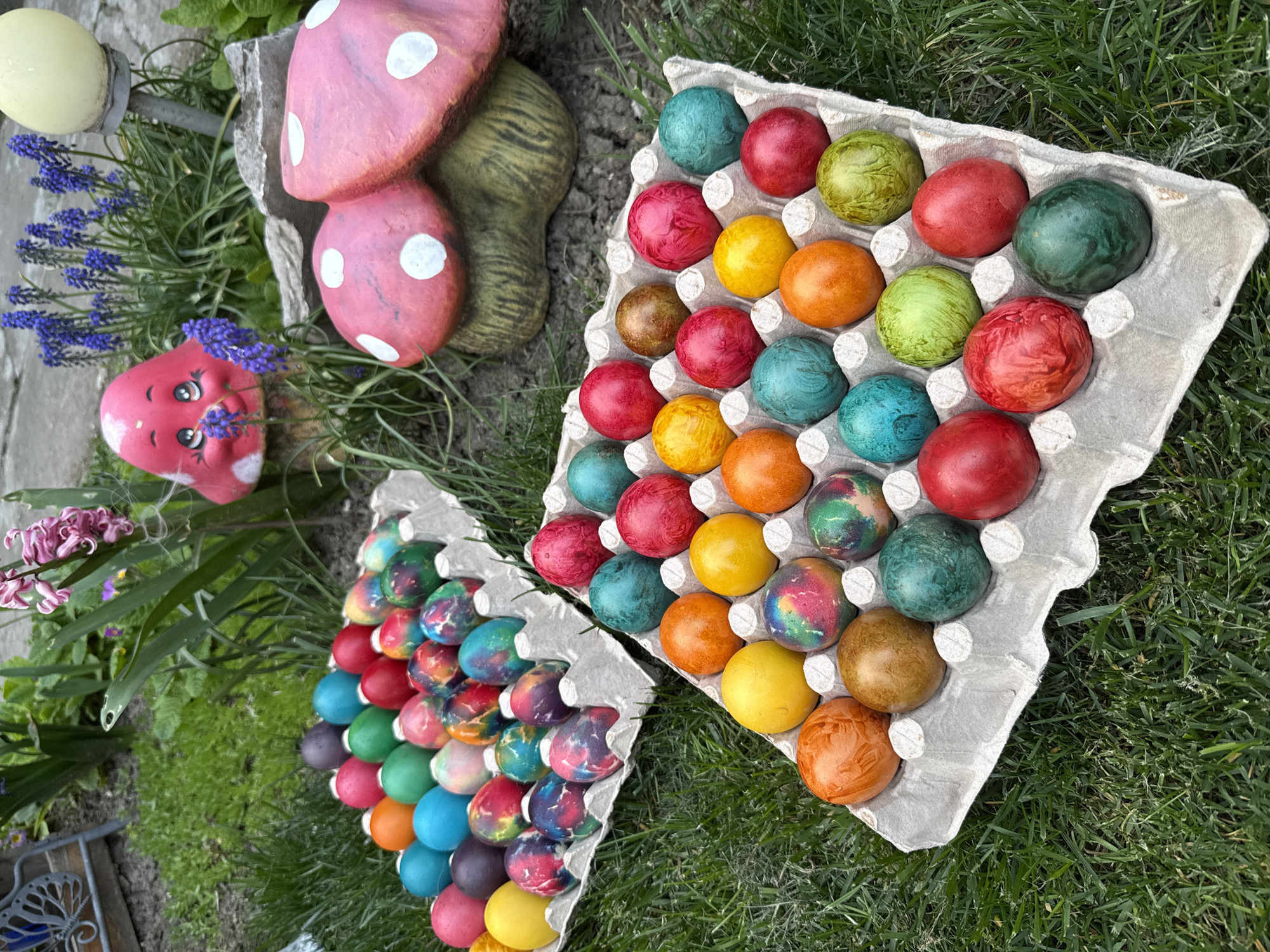 The image depicts a garden scene with two partially-filled egg cartons containing dyed Easter eggs. One carton is relatively full with eggs in various solid colors (red, blue, green, yellow, orange, brown) and patterns. The other carton is partially filled with eggs also displaying various colors and patterns, including tie-dye effects. The cartons are placed on green grass. Adjacent to the cartons, there are garden decorations, including red mushroom figurines with white polka dots, a yellow orb on a stake, a red face figurine, and some purple flowers. The location is likely a domestic garden setting, potentially in Lukovit, Bulgaria.