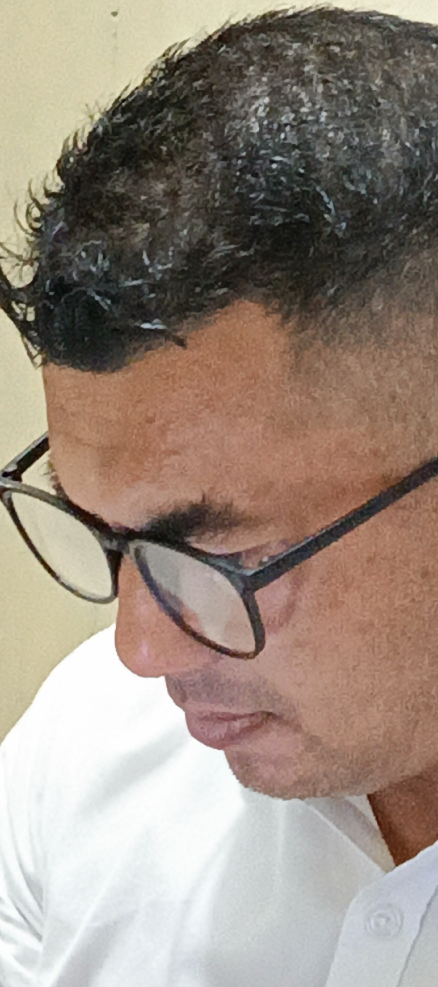 The image is a close-up shot of a person wearing glasses and a white shirt. The person has dark, curly hair and is looking downwards. The background is a plain, light-colored wall. The lighting appears to be from above, casting shadows on the face. The image does not contain any specific elements that would allow for identification of the location within Banda Aceh, Indonesia.