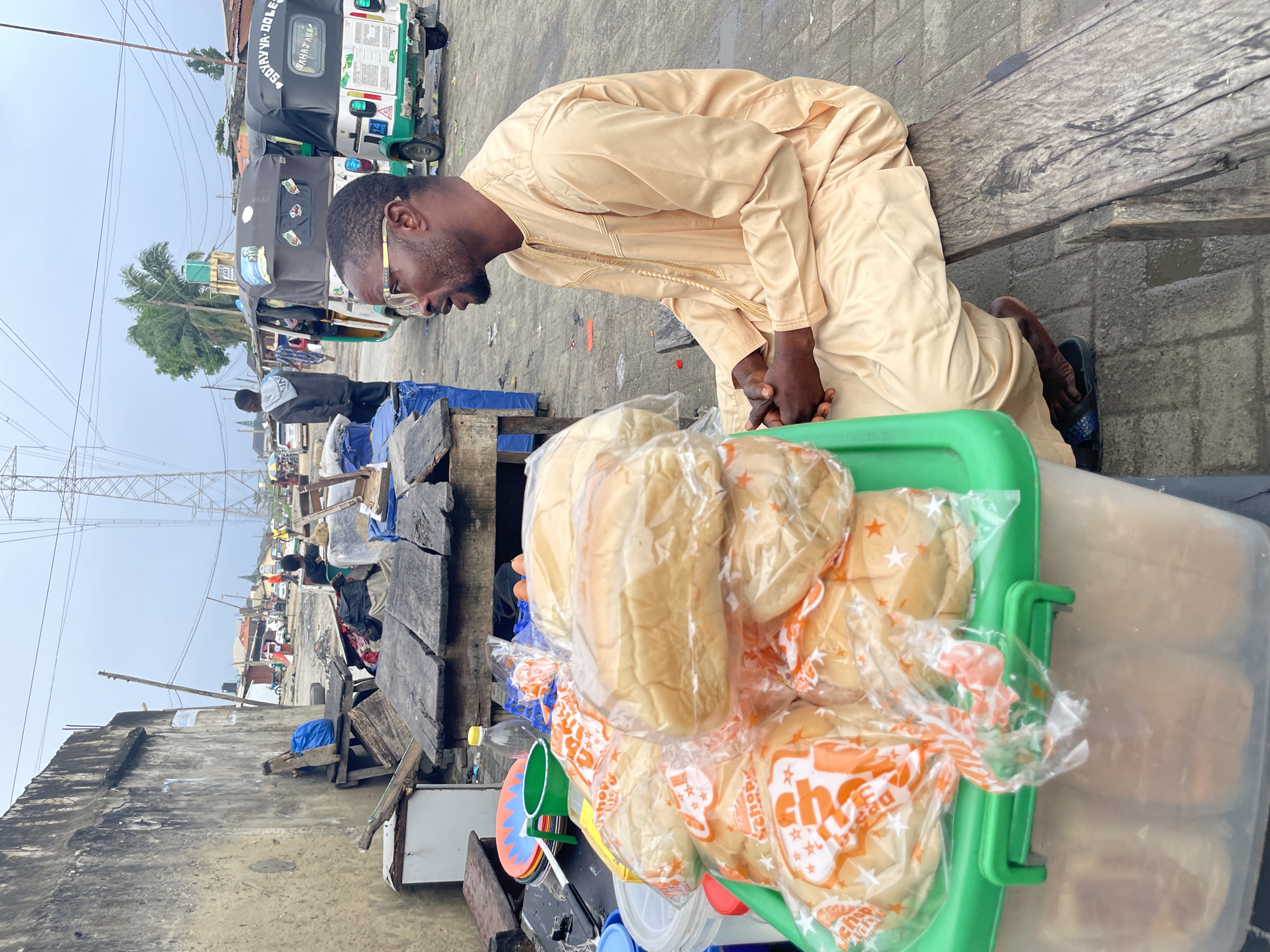 The image depicts a roadside scene in the city of Ado, Nigeria, under an overcast sky. In the foreground, a dark-skinned man with a beard and eyeglasses, wearing a light-colored (beige/cream) traditional garment with gold embroidery, sits in profile on a rough wooden bench, facing left. His hands are clasped, and he wears dark sandals. To his immediate left, a green plastic container holds multiple clear plastic bags filled with bread rolls, each bag featuring orange branding that reads "Chop Life Bread" and star motifs. Adjacent to the bread, on a rudimentary wooden table, are various items including stacked colorful plastic plates, a green plastic mug, a clear plastic bottle, and a blue plastic egg tray containing eggs. A white refrigerator unit is partially visible on the far left.
The midground reveals a cluttered street environment. Behind the sitting man and the vendor's setup, several dilapidated wooden tables and structures are present. A person in a dark hoodie stands further back on the left. On the paved street, two auto-rickshaws (*keke napep*) are visible. The one on the right is green and white, displaying "SOYAYYA-DOLE" and "MAHATABA" in white text on its rear, along with a license plate "141 HHE". The other *keke napep* is predominantly dark-colored. Other individuals are dispersed along the street. The buildings lining the street are mostly single or two-story structures, some with corrugated iron roofs, indicating a typical urban or semi-urban setting. A tall palm tree is visible in the mid-distance.
In the background, a large, metallic lattice power transmission tower dominates the right side, extending high into the uniformly cloudy sky. Overhead power lines crisscross the frame. The ground surface varies from paved concrete, showing wet patches, to unpaved or dirt sections, with scattered debris. A weathered, discolored concrete wall with visible cracks forms the left boundary of the scene.
