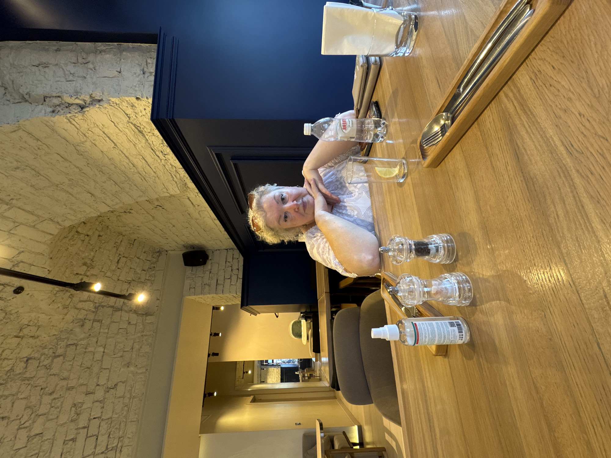 An overhead shot captures a woman seated at a wooden table in what appears to be a restaurant or cafe. The woman, with blonde hair and wearing a white top, rests her chin on her clasped hands. In front of her on the table are a bottle of water, a glass with a lemon slice, a salt and pepper shaker set, and a bottle of hand sanitizer. To the right, a napkin rests in a glass. The background reveals a brick-lined arched ceiling and dark blue walls, suggesting an indoor setting. The establishment is identified as being in Kazan, Russia.