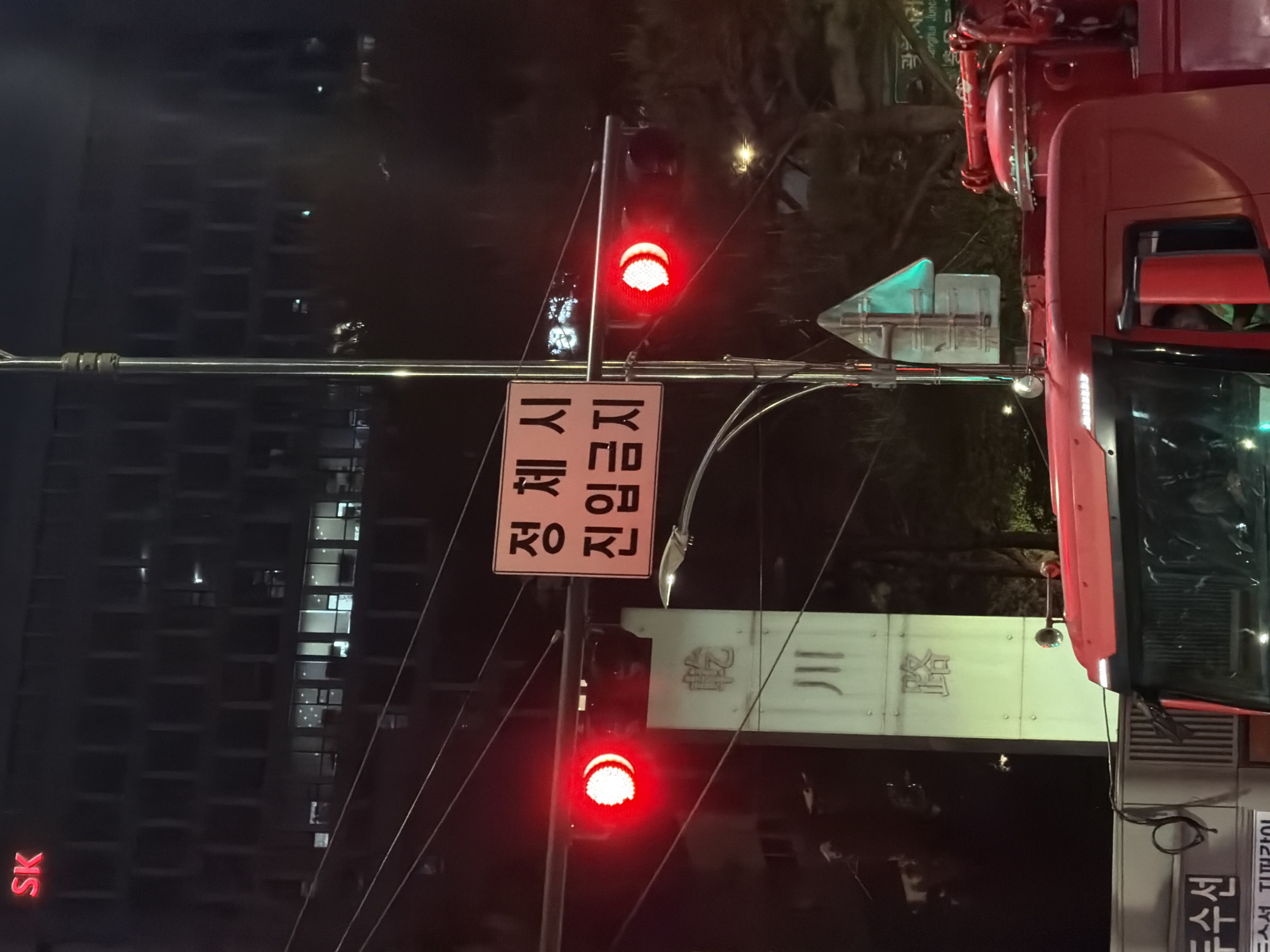 The image shows a street scene in Seoul, South Korea at night. A red traffic light is illuminated above a rectangular white sign with Korean text. A building with the SK logo is visible in the background. A red vehicle, possibly a firetruck, is in the right portion of the frame. A green street sign and white rectangular sign with Korean text, possibly designating a road or a neighborhood, are also visible. The image is slightly rotated counterclockwise.