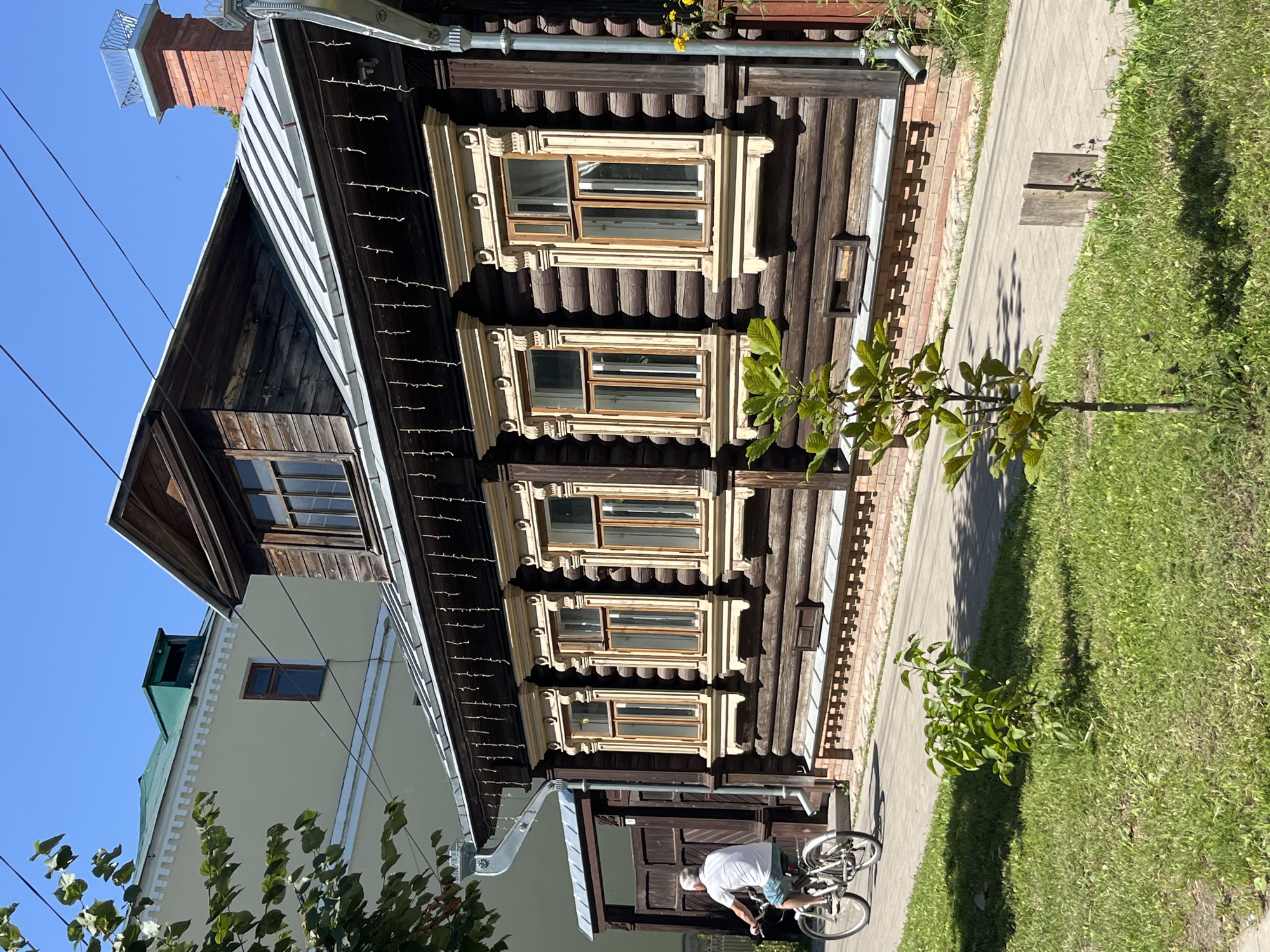In Kladovitsy, Russia, a man cycles past a historic wooden house on a sunny day. The house, constructed of dark logs, features ornate window frames with light-colored trim and decorative carvings. String lights hang along the eaves, adding a festive touch. A metal roof slopes upwards towards a dormer window. The house is bordered by a brick foundation and a paved walkway. To the left, a modern light-yellow building with a green roof peeks out from behind lush green foliage. The ground is covered in green grass, with a young tree growing in the foreground. The sky is a clear blue.