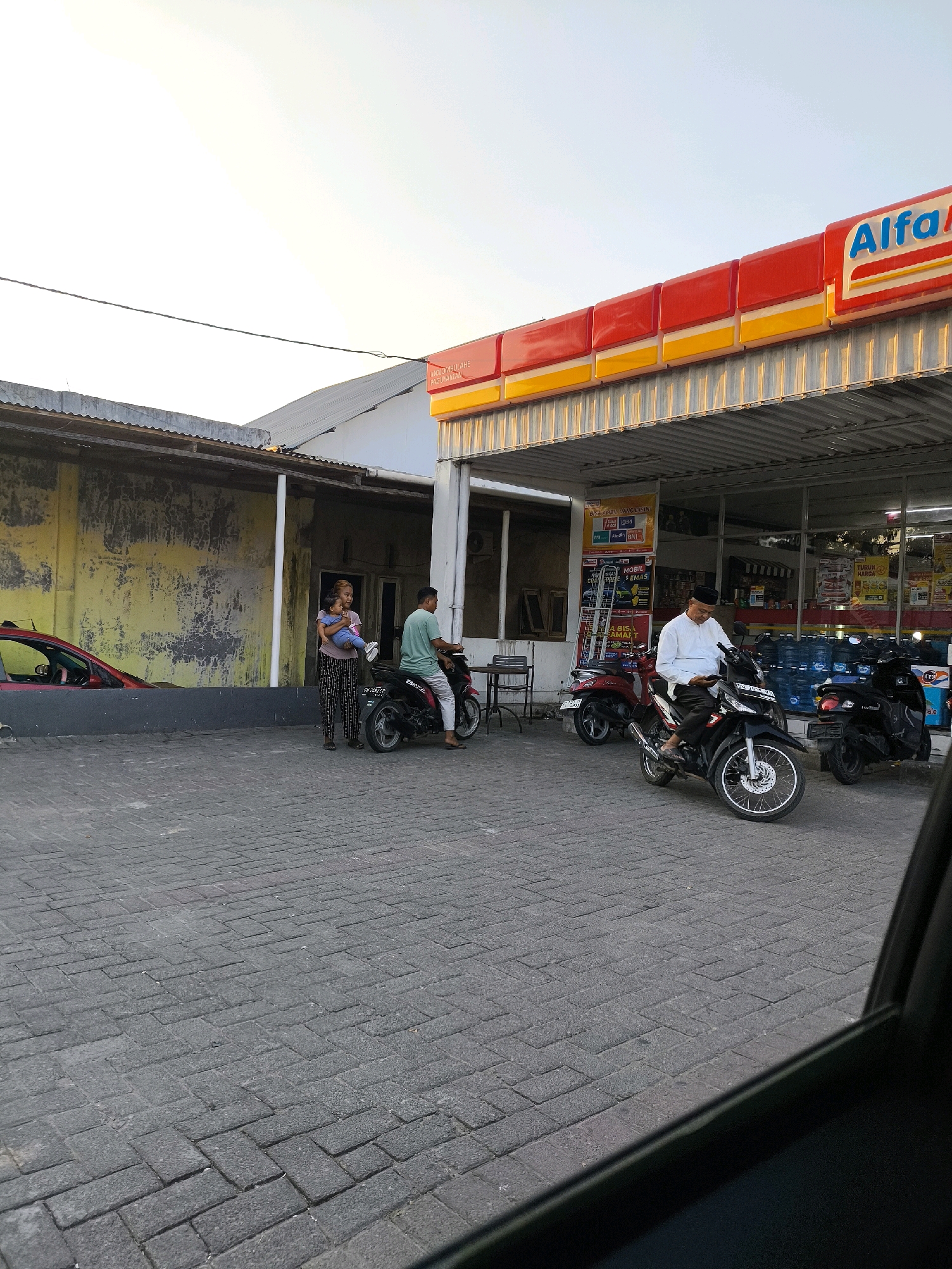 In Tenilo, Indonesia, a street scene unfolds in front of an Alfamart convenience store. The paved ground, composed of interlocking grey bricks, stretches across the foreground. To the left, a red car is partially visible. Towards the center of the image, two women stand near a parked motorcycle, one holding a child. A man sits on a black motorcycle to the right of them, facing a storefront. Behind him, another man in a white shirt and a peci hat is seated on a motorcycle, looking at his phone. The Alfamart store features a prominent red and yellow awning and glass doors revealing shelves stocked with products. Several other motorcycles are parked in front of the store. The sky above is clear and bright, indicating daylight.
