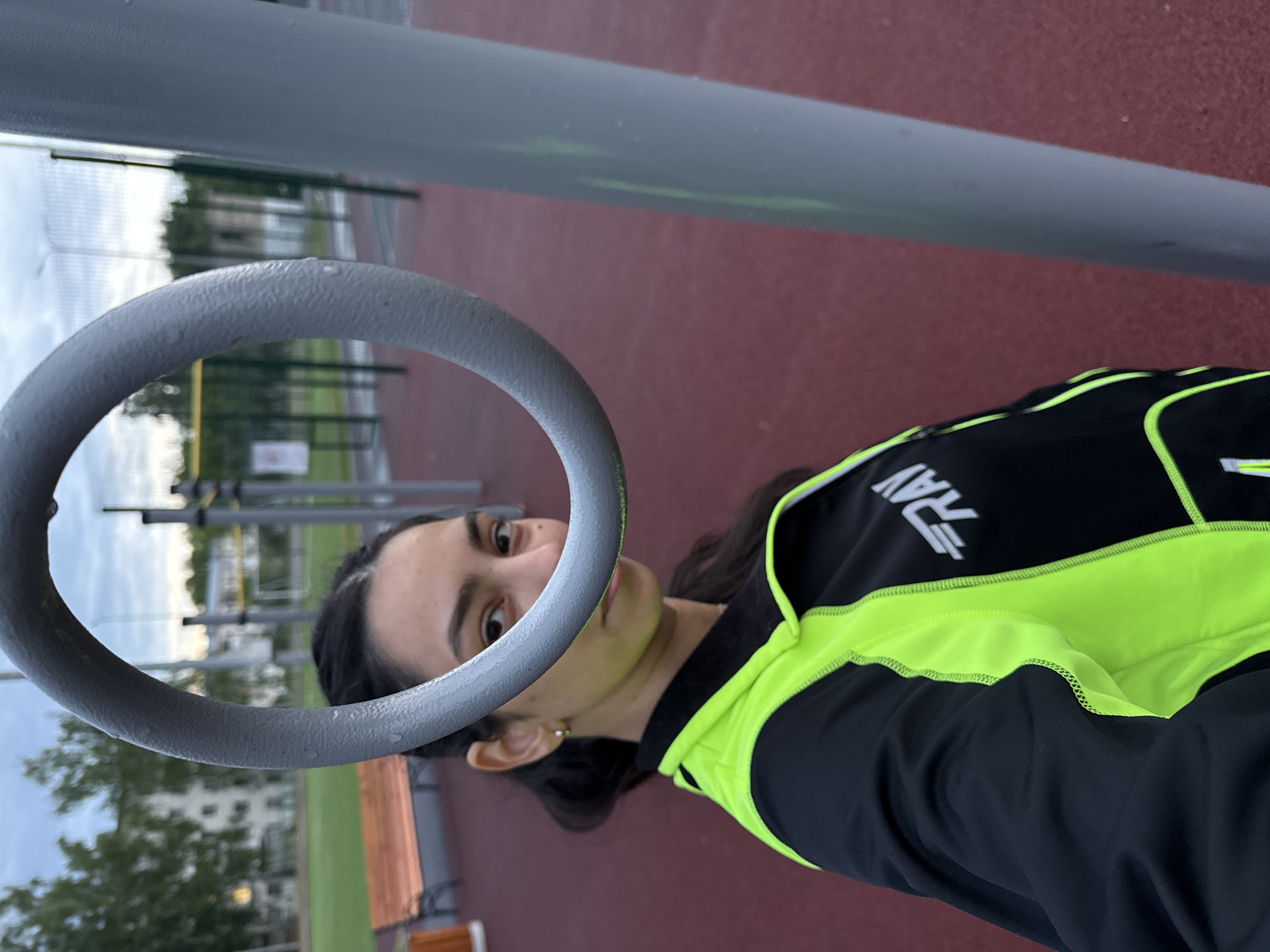 A person with dark hair, wearing a black and neon green athletic jacket, is visible through a grey metal ring. The person's face is partially visible, with one eye clearly seen and a neutral expression. The background features outdoor fitness equipment, including parallel bars and other metal structures, located on a red athletic surface. The location appears to be a park in a city, evidenced by trees, buildings, and open sky visible in the background. Given the prompt, the location is Chaikovsky, Russia. The person is likely at an outdoor fitness area in the city.