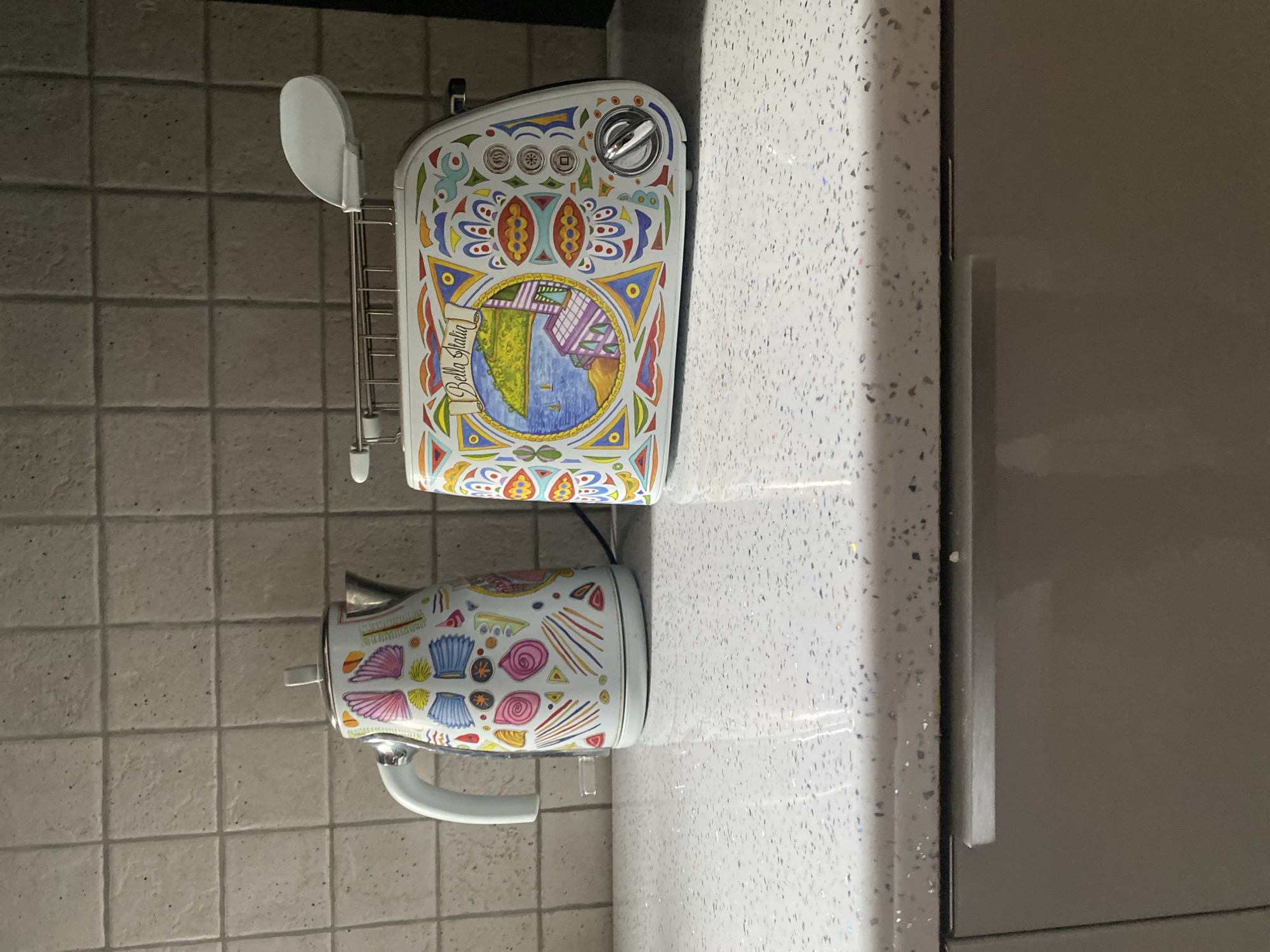The image shows a white kettle and a white toaster, both decorated with colorful, intricate patterns. The kettle has a white handle and a silver lid. The toaster has a metal rack on top and various buttons and a dial on its side. Both appliances are placed on a speckled white countertop. Behind them, a tiled backsplash with beige square tiles is visible. The lower part of the image shows the top of a grey cabinet with a horizontal silver handle. The scene is indoors, likely a kitchen. There is no indication of the city of Wrocław, Poland.