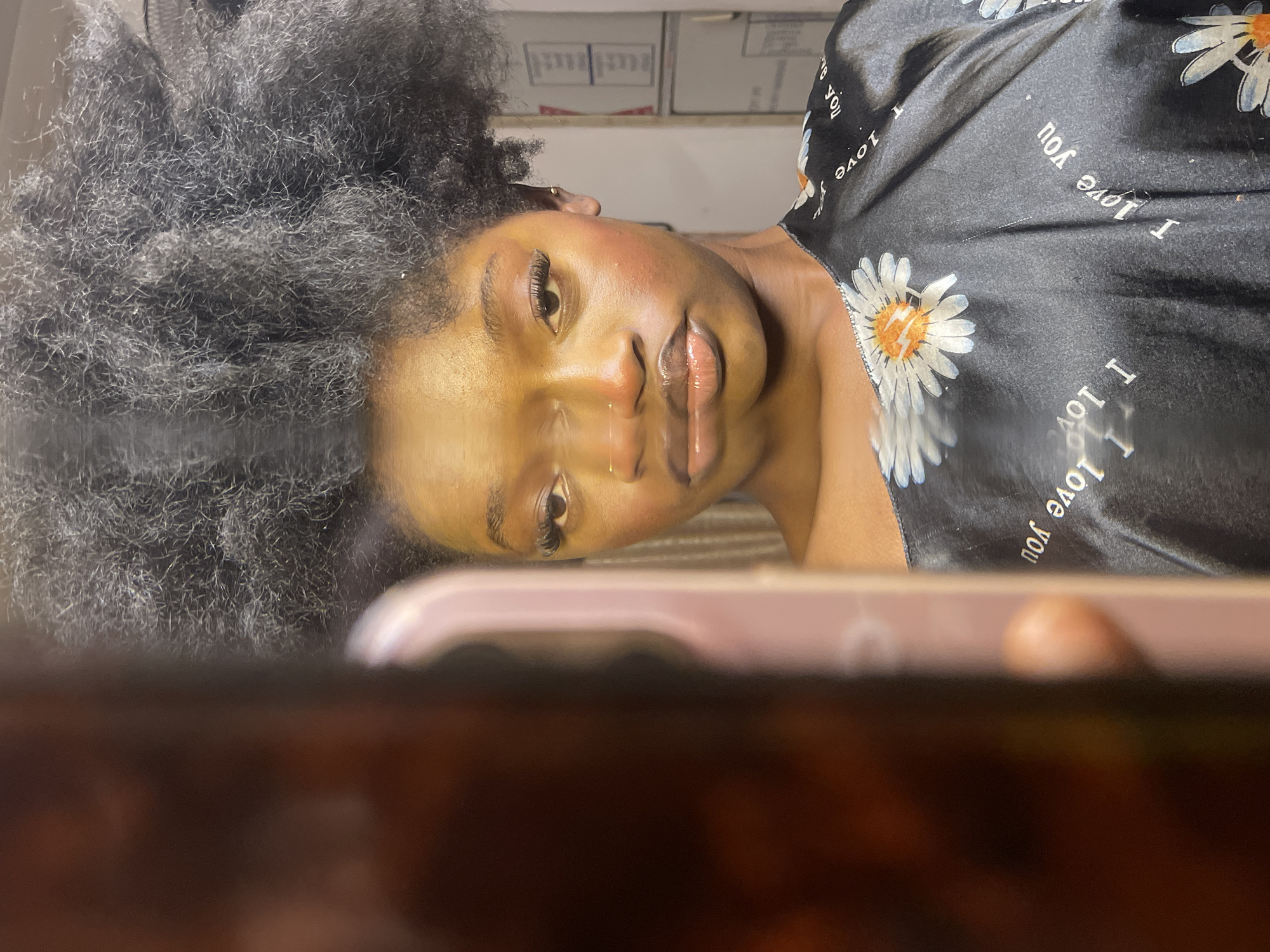 A woman is seen from the chest up, facing forward with her head slightly tilted to her left. She has dark, voluminous curly hair styled upwards. Her skin is dark, and she is wearing makeup, including noticeable false eyelashes and a glossy lipstick. She has a nose ring. Her top is black with a daisy pattern and the words "I love you" printed on it. She is looking directly at the camera, which appears to be a phone camera, creating a reflection effect. The background is out of focus and appears to be an indoor setting, possibly a room with boxes stacked against a wall. The lighting suggests an indoor environment. The image does not provide any specific details that would allow for identification of a location within Ado, Nigeria.