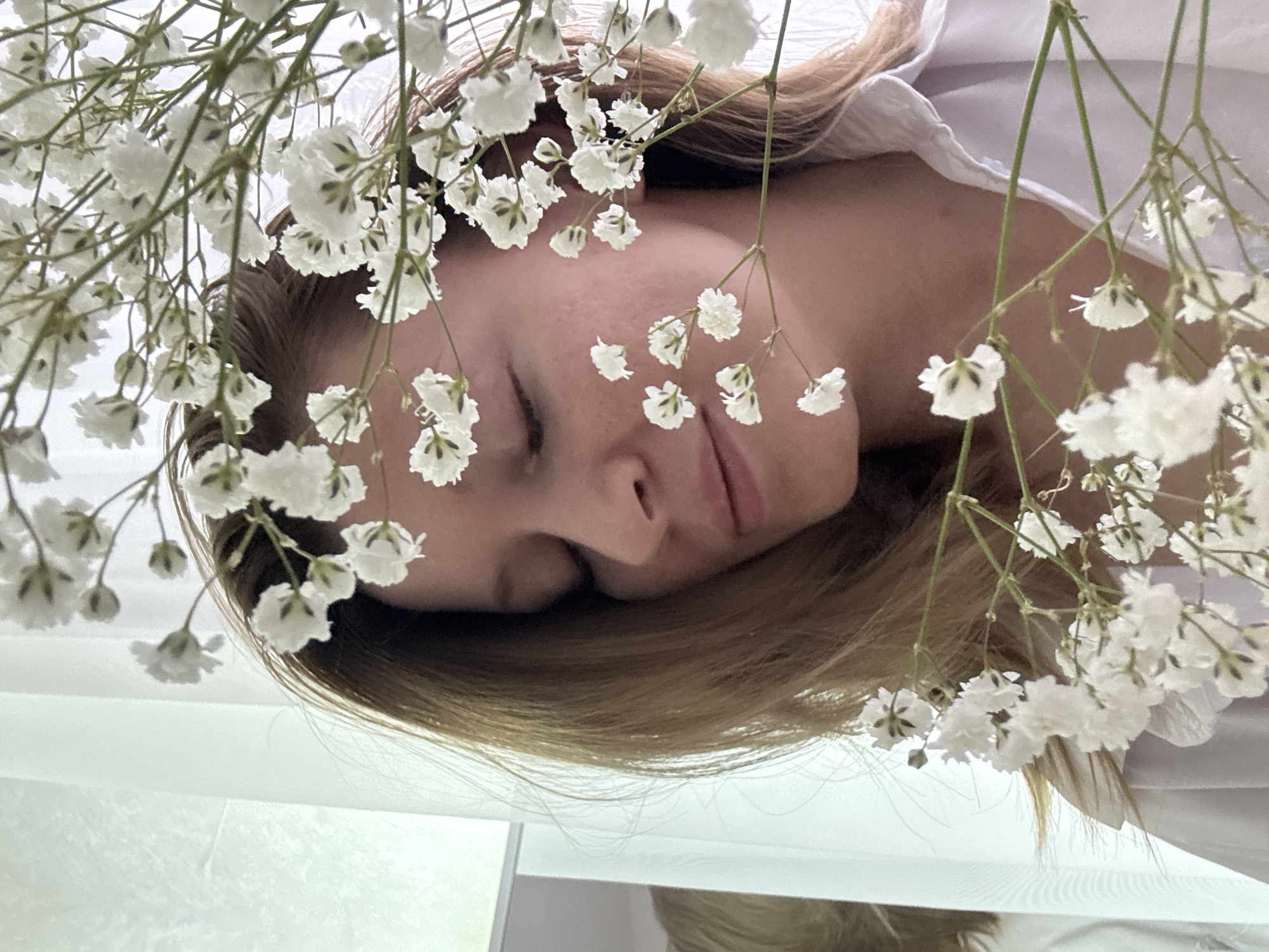 A woman with fair skin and light brown hair is lying down, surrounded by sprigs of white baby's breath flowers. She is wearing a white shirt. The background is a blurry white surface. There is no information available to confirm the location as Chaikovsky, Russia.