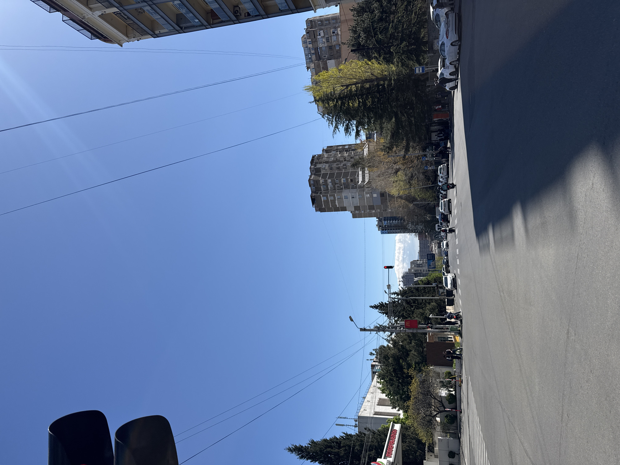The image shows a street in Tbilisi, Georgia. The street is flanked by buildings and trees, with a clear blue sky above. Cars are parked along the side of the road, and a few people and motorcycles are visible on the street. A traffic light is showing red. A McDonald's sign is also visible.