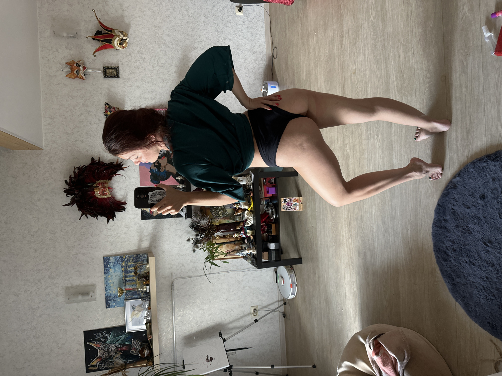 A woman is posing for a selfie in a room that appears to be a living space, possibly in Vladimir, Russia. She is wearing a dark green top and black underwear. Her body is positioned to highlight her legs, with one leg bent and the other extended. She is holding a smartphone in her right hand to take the picture.
The room has a light-colored, textured wallpaper on the walls and a wooden floor. Several decorative items are visible, including Venetian masks on the wall, a collection of figurines on a black shelf, and several framed pictures or artwork. A robot vacuum cleaner is on the floor near the shelf, and an easel with a canvas is also visible in the lower part of the frame.
The lighting suggests it might be daytime, with natural light casting shadows on the floor. The overall mood seems relaxed and perhaps playful, given the selfie pose. There is no visible text in the image.