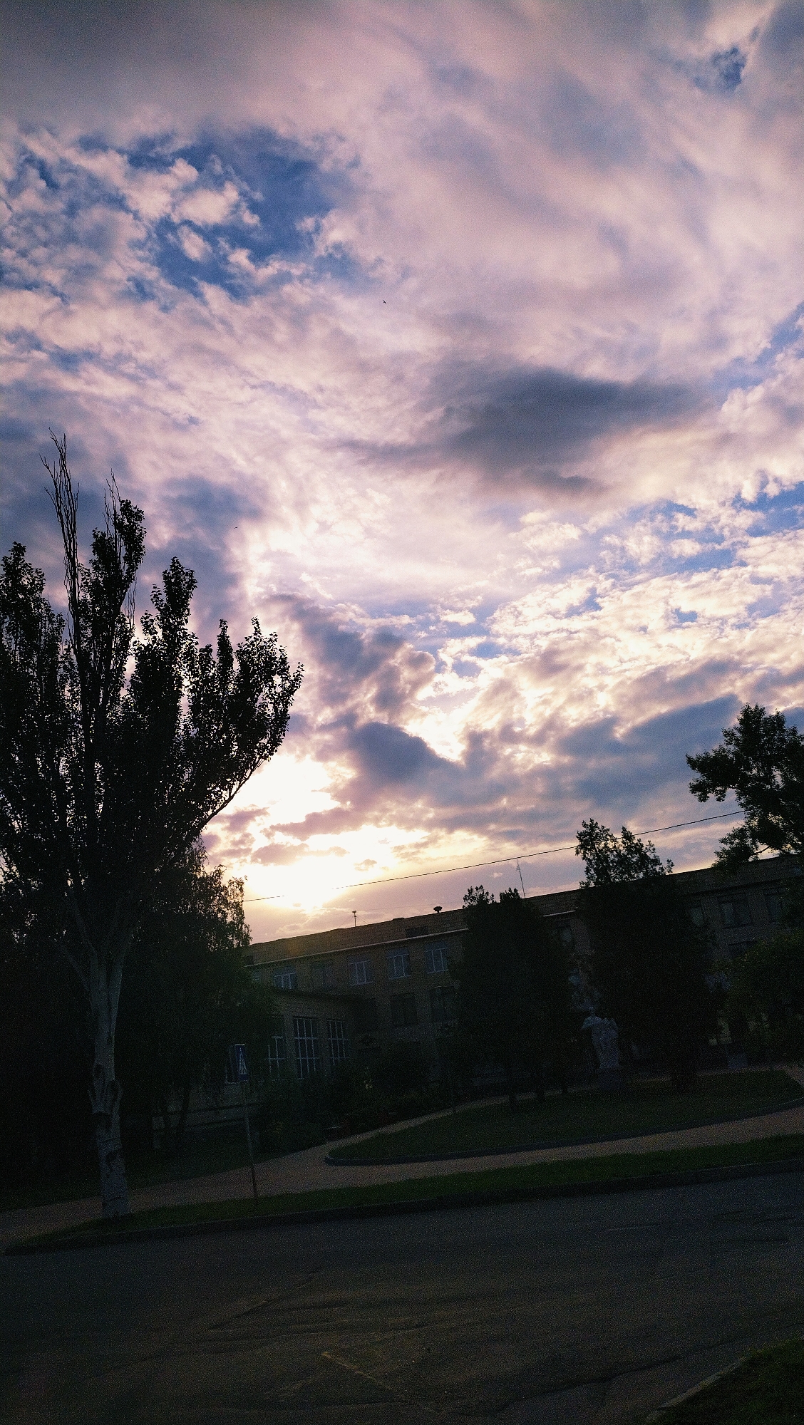 The image depicts a serene sunset scene in Horishni Plavni, Ukraine. The sky is adorned with a dramatic display of clouds, painted in shades of purple, pink, and grey, with patches of bright blue peeking through. The setting sun casts a warm glow, illuminating the undersides of the clouds and creating a soft light that bathes the entire scene.
In the foreground, a dark, silhouetted tree stands prominently on the left side of the frame. Its branches reach upwards, silhouetted against the vibrant sky. To the right of the tree, a multi-story building with a light-colored facade is visible, its windows reflecting the sky's colors. Trees line the front of the building, adding a touch of greenery to the scene. A paved area or road runs across the bottom of the image, appearing dark and somewhat worn, suggesting it might be an asphalt surface. The overall atmosphere is peaceful and tranquil, with the natural beauty of the sunset dominating the composition.