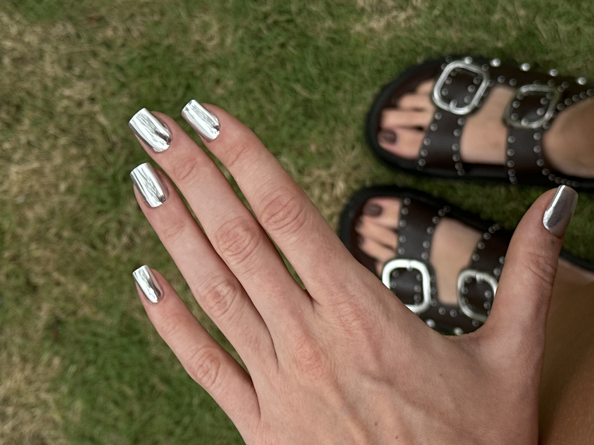 A woman's hand with metallic silver nail polish is displayed against a backdrop of green grass. Her foot is visible in the frame, clad in brown leather sandals with silver buckles and studs. The location is presumed to be outdoors, given the presence of grass. There is no evidence to support the location being in Dalung, Indonesia.