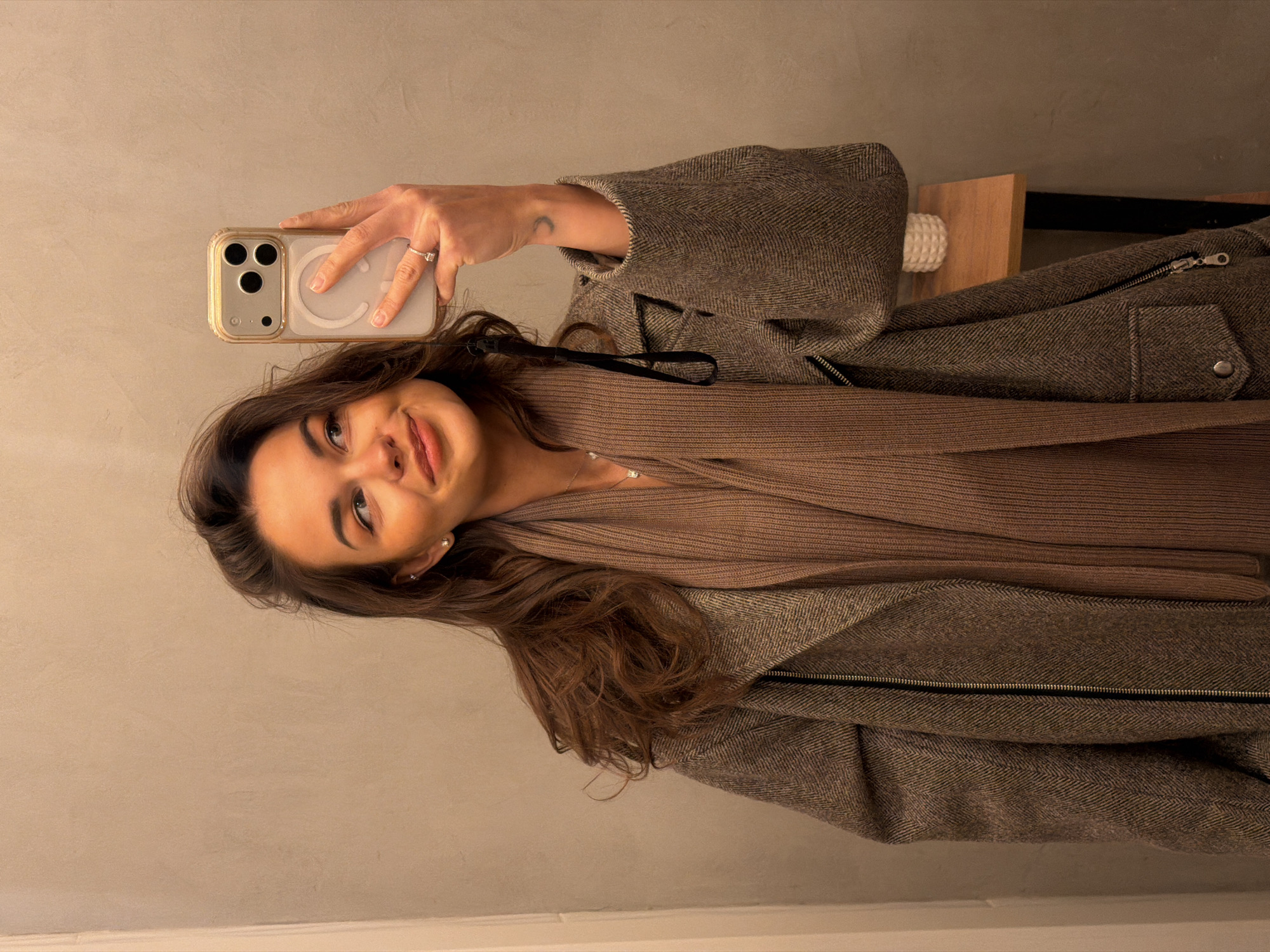 A woman stands indoors, facing left, taking a selfie with her iPhone. She's wearing a brown wool coat over a brown knitted top or scarf. Her hair is long, brown, and wavy. She has fair skin and is wearing a ring on her left hand. The phone has a clear case with a MagSafe circle. Behind her, there is a light-colored wall and a piece of wooden furniture. There is no information in the image to suggest the location is Krasnoyarsk, Russia.