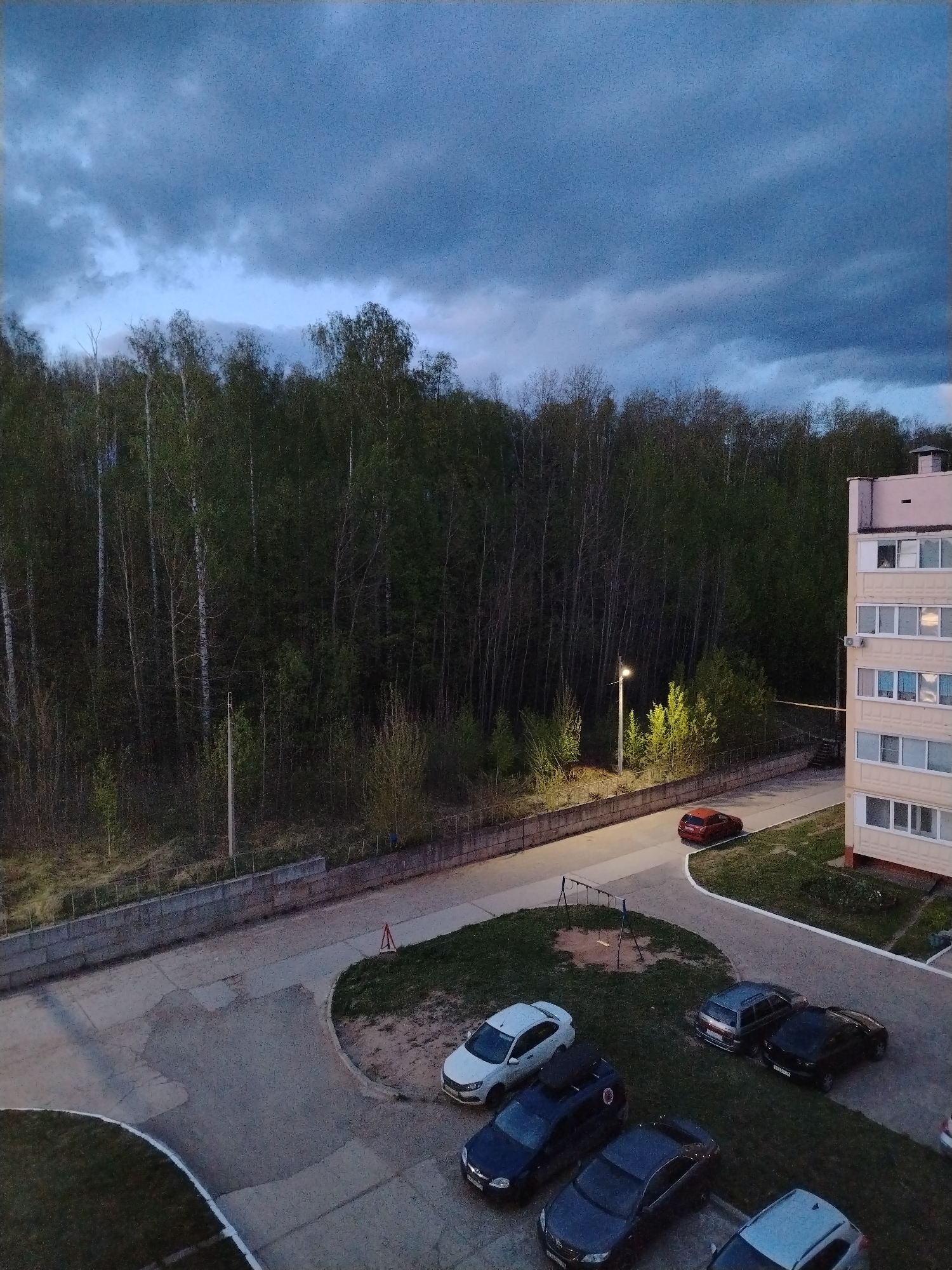 The image depicts an outdoor scene in Noviy, Russia, at twilight or dusk. The scene is captured from an elevated position, possibly from a building overlooking a parking area and surrounding environment. The sky is overcast with dark, heavy clouds.
Adjacent to the parking area is a dense forest with a mix of deciduous and evergreen trees. The trees are bare or have only begun to leaf out, suggesting it is early spring. A concrete retaining wall separates the parking area from the forested area.
A street with a single lamppost illuminates a portion of the road leading away from the parking area.
The parking area contains multiple parked cars, including white, blue, gray, and black vehicles. The parking surface appears to be concrete or asphalt. A small playground with swings is visible on a grassy patch adjacent to the parked cars. A residential building is on the right side of the image with multiple windows.