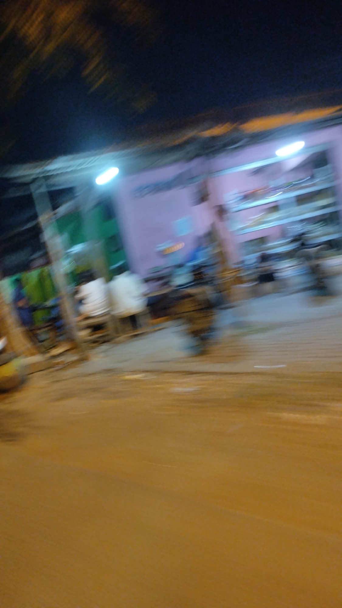 The image depicts a blurry, nighttime street scene in Sambi Bulu, Indonesia. A building with a pink facade and a roofed overhang is visible on the right. Shelves are mounted on the interior wall of the building, suggesting it may be a shop or restaurant. Several individuals, largely indistinct due to motion blur, are seated at tables or standing. One person is seated on the left, facing away from the camera, wearing a white shirt. Another individual in a white shirt is seated to their right. Further to the right, more figures are present, some appearing to be on motorcycles or scooters. The street in front of the building is paved, and the area is illuminated by artificial lights, casting a warm glow. The sky above is dark, with streaks of orange light possibly from distant streetlights or vehicles. The overall impression is one of a lively, urban setting at night.
