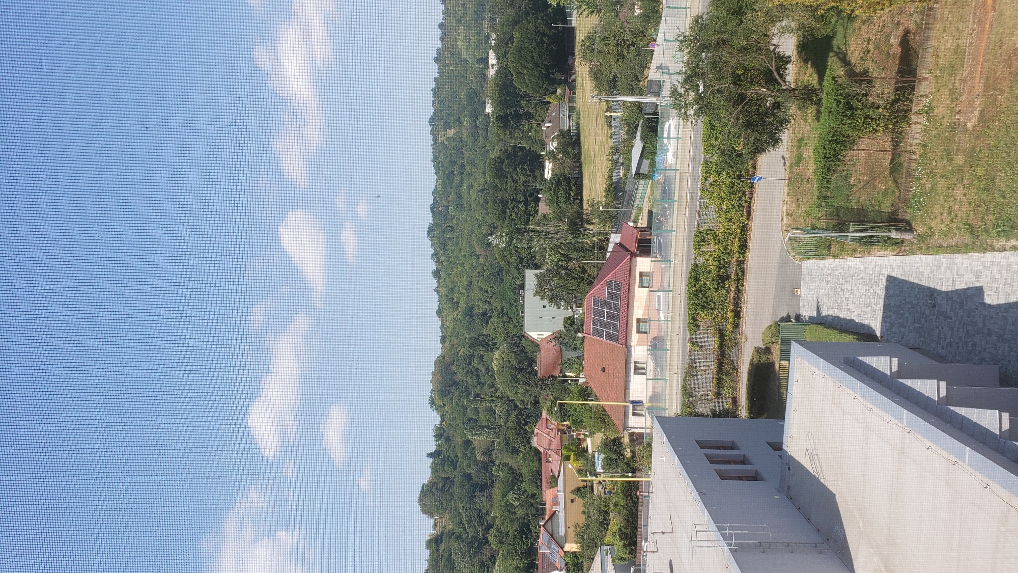 The image shows a residential area with a view of a wooded hillside. In the foreground, there is a modern grey building with a flat roof. A paved walkway leads away from the building towards a street. Beyond the street, there are several houses with red tile roofs, some of which have solar panels installed. The houses are surrounded by greenery, and a dense forest covers the hillside in the background. The sky is blue with a few scattered white clouds. The image appears to be taken from a window with a screen. There are no people visible in the image.