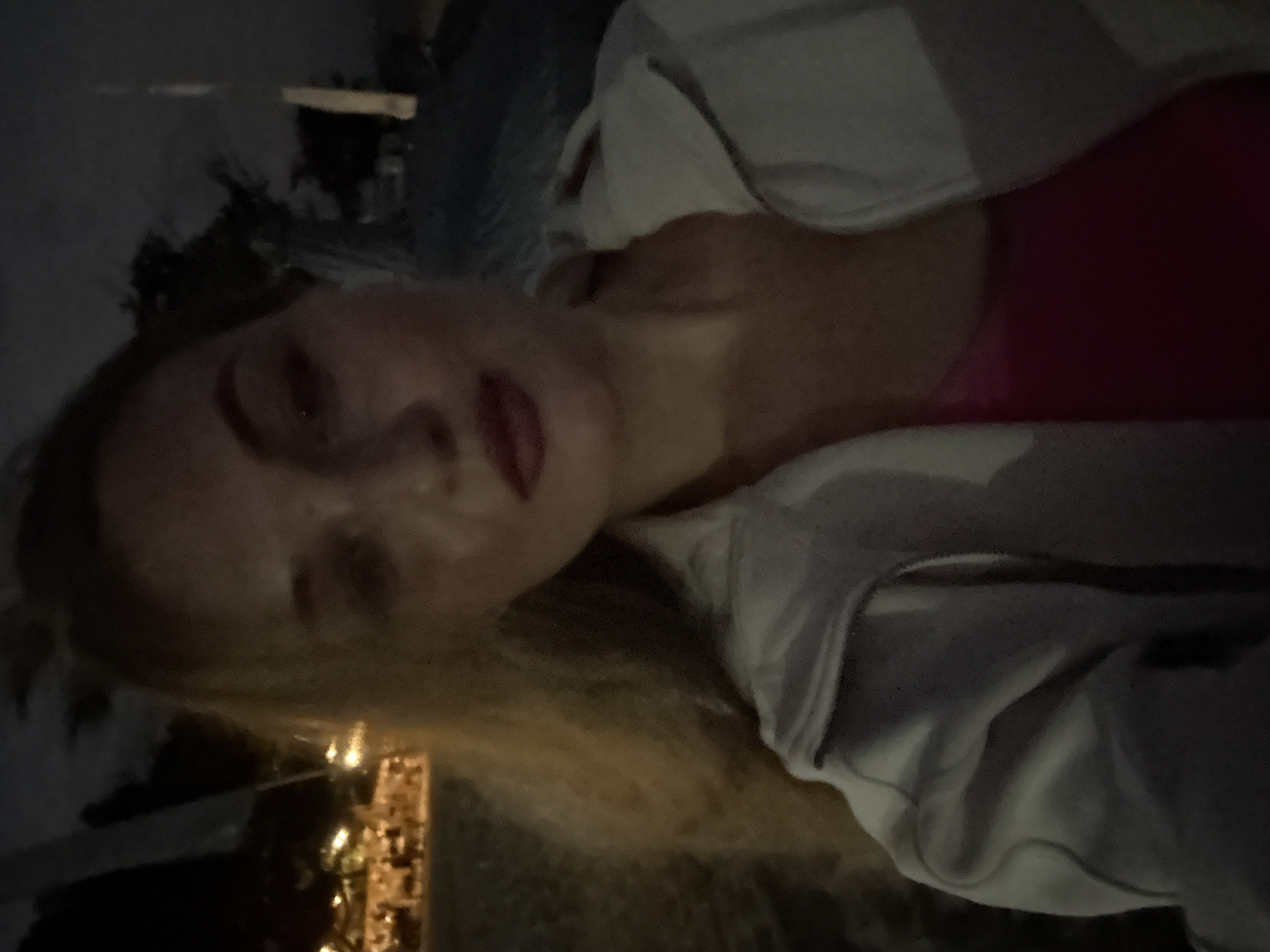 An extreme close-up, low-light selfie shows a woman with long blonde hair and red lipstick looking at the camera. She is wearing a light-colored zip-up jacket over a pink top. The background is dark and blurry, with some indistinct lights that suggest an outdoor setting at night. The setting is identified as a street in Denpasar, Indonesia, possibly a beach area or a busy street lined with shops or restaurants.
