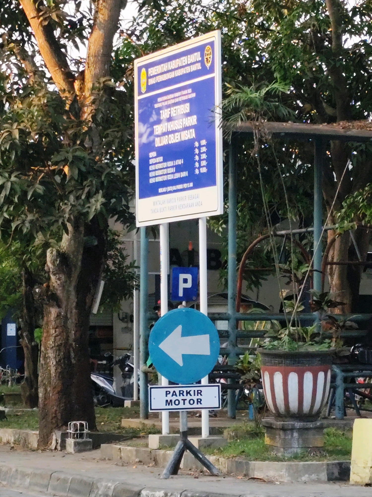 This is a medium shot, eye-level photograph taken outdoors in Bantul, Indonesia. The scene features a tall, blue and white sign with text and a yellow emblem at the top. The sign lists "PEMERINTAH KABUPATEN BANTUL" (Bantul Regency Government) and "DINAS PERHUBUNGAN KABUPATEN BANTUL" (Bantul Regency Transportation Agency). Below this, it indicates "TARIF RETRIBUSI" (Fee Schedule) for "TEMPAT KHUSUS PARKIR DILUAR OBJEK WISATA" (Special Parking Area Outside Tourist Attractions). It then lists prices for different vehicle types. A smaller white sign with the word "PARKIR" (Parking) and "MOTOR" (Motorcycle) is mounted below a blue circular sign with a white left-facing arrow.
In the background, trees with dense green foliage are visible, along with portions of a building or structure, possibly a covered area with metal railings. A large decorative pot with red and white vertical stripes stands to the right, filled with plants. To the left of the sign, a portion of a tree trunk is prominently featured. In the middle ground, a motorcycle is parked. The ground is paved with concrete and there is grass and dirt. The sky is not visible. The overall lighting suggests it may be late afternoon or early morning, with sunlight illuminating parts of the trees and signage.