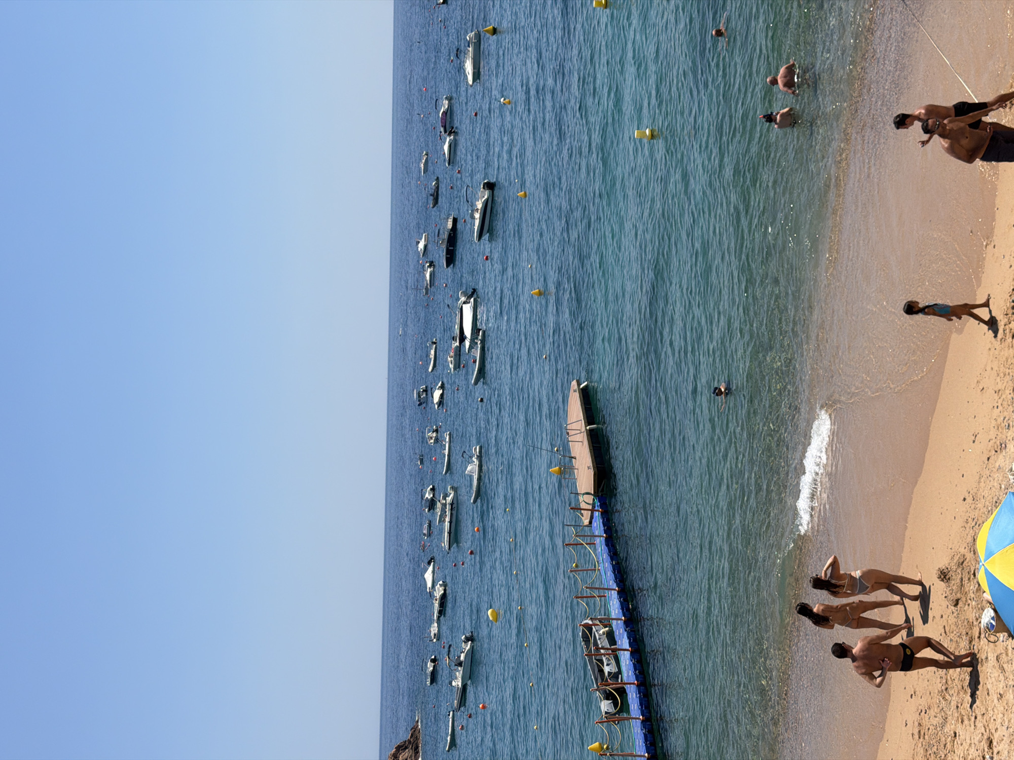 This image depicts a sunlit day at a beach in Tossa de Mar, Spain. The scene is divided between a sandy beach in the foreground and a calm, blue sea filled with numerous boats in the background.
On the beach, several individuals are visible. A man in dark swim trunks stands on the sand, facing away from the viewer. Two women in bikinis walk towards the water. Further down the beach, a young girl in a blue swimsuit walks along the shoreline. To the right, two men, one appearing to be speaking, are standing near the water's edge. In the sea, several people are swimming or wading, including a lone swimmer in the upper right section of the water.
The sea is dotted with a variety of boats, mostly smaller motorboats, anchored or drifting. A blue floating dock with a wooden platform extends into the water from the left side of the image, with a small dinghy tied to it. Yellow buoys are scattered throughout the water, likely marking swimming or boating areas. The water is clear, allowing a view of the seabed in some shallower areas. The sky is a clear, bright blue, indicating fair weather. The overall atmosphere is one of a relaxed summer day at a Mediterranean coastal resort.