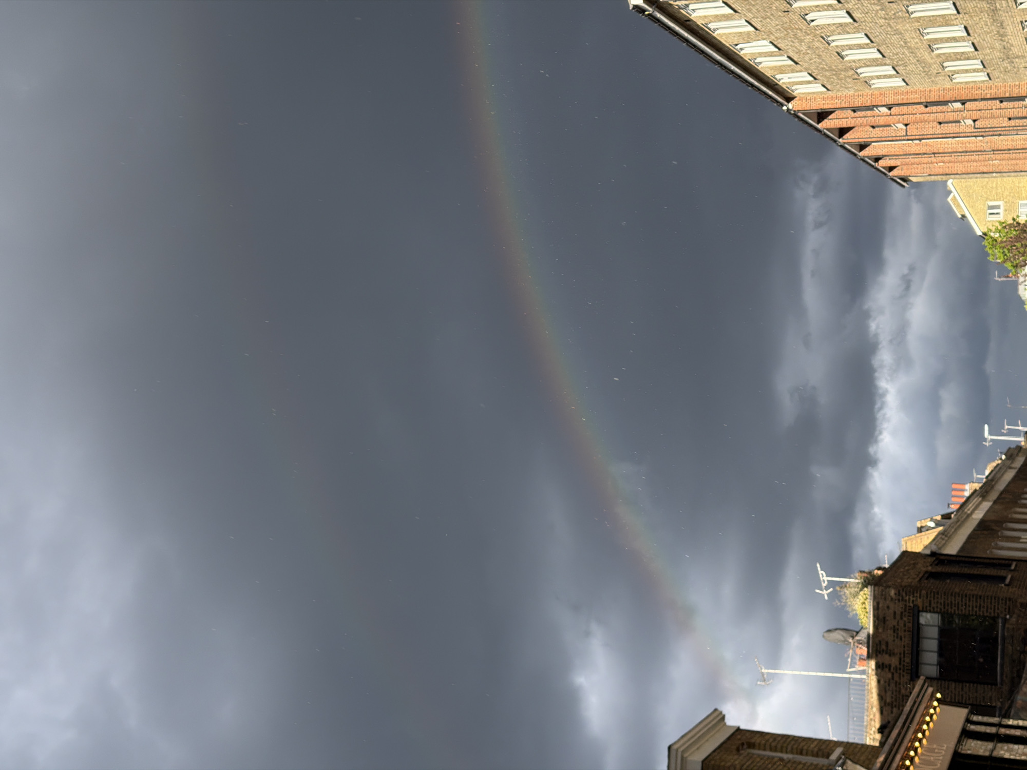 The image depicts a rainbow in a cloudy sky between buildings in London, United Kingdom. The buildings are brick and stone, and the sky is dark grey. No people are visible.