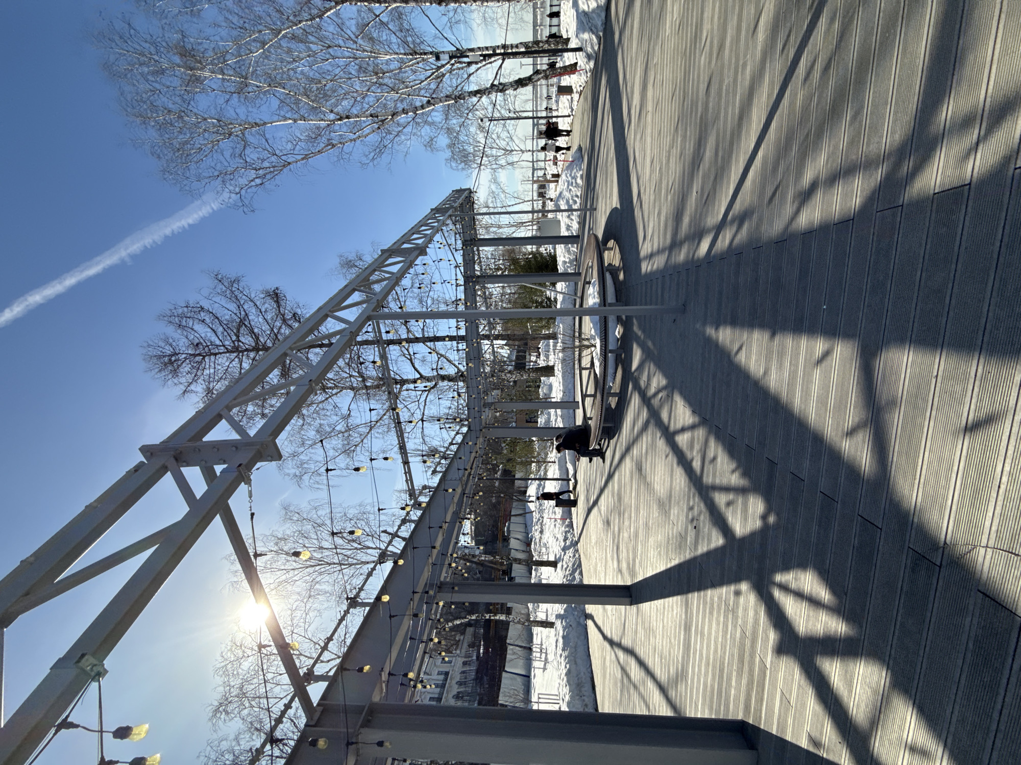 The image captures a wide view of the Perm Embankment along the Kama River in Perm, Russia, on a bright, sunny winter or early spring day. The foreground and midground feature a broad wooden plank walkway composed of light-colored slats.
A large, open metal framework structure, likely a pergola, dominates the left and central portions of the scene. This structure consists of gray metal beams and columns, supporting multiple strands of string lights with exposed bulbs. Strong sunlight from the upper left casts elongated, angular shadows of the metal framework and columns across the wooden walkway.
To the left of the walkway, the ground is covered in snow, and bare deciduous trees are visible. A multi-story building with numerous windows is partially discernible in the distant background, behind the metal structure, partially obscured by a white fence and a dark barrier.
In the mid-ground, beneath the metal structure, one individual wearing dark clothing is seated on a curved wooden bench. Further along the pathway, another individual is walking away from the camera, passing a dark cylindrical object. Several other figures are visible walking in the distance along the embankment pathway to the right.
The right side of the image features snow-covered ground and numerous bare deciduous trees, including several prominent birches, lining the edge of the walkway. Beyond the trees, a large body of water, the Kama River, is visible in the distant background. The sky is clear and blue, with a distinct white aircraft contrail visible in the upper right quadrant.