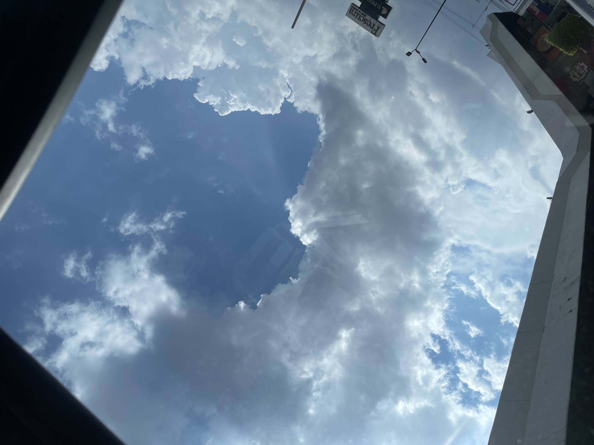 The image, taken from a low angle, depicts a partially cloudy sky above the city of Puebla, Mexico. A section of clear blue sky is visible amidst a field of cumulus clouds. The camera is likely positioned indoors looking upwards, as evidenced by the frame of a window or sunroof visible along the edges of the image. The perspective suggests the photograph was taken from within a car or building. The environment appears to be urban, with signage of "Frescura" visible, and elements of buildings in the periphery.