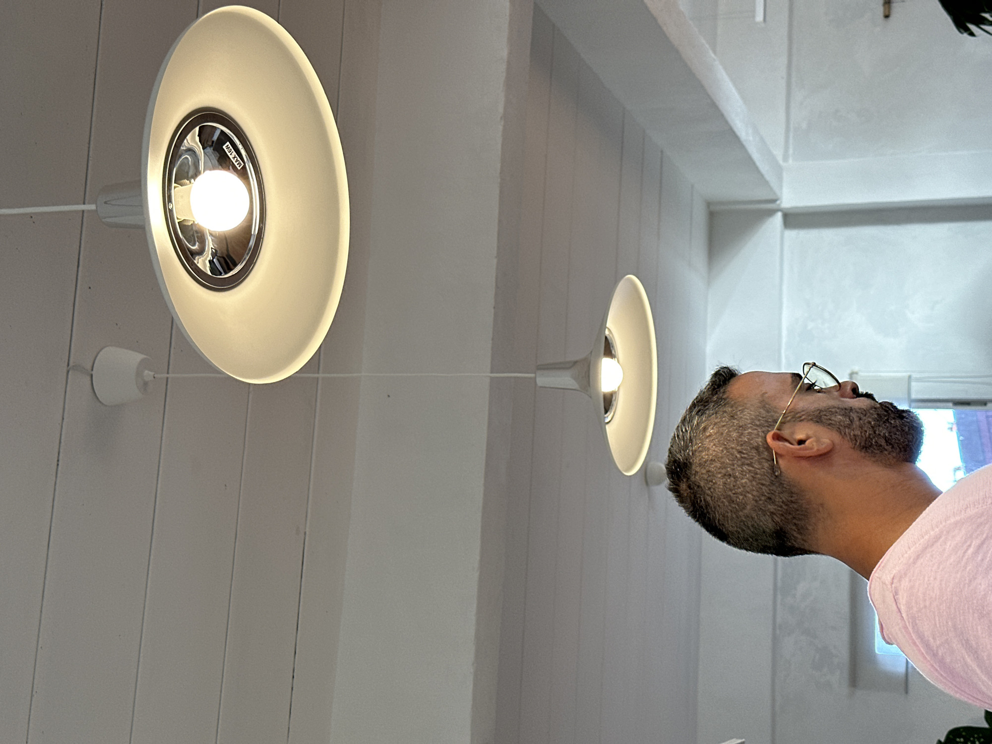 The image shows an indoor scene with a man wearing glasses and a pink shirt in the foreground, looking down and to the right. In the background, two white pendant lights hang from a white slatted ceiling. The man's hair is short and grey, and he has a beard. One light fixture is closer and brighter, showing a label that reads "MAX 15W". The overall environment appears to be a clean, modern space, possibly a cafe or a restaurant. The location is specified as Koh Phangan, Thailand, but the image itself does not contain specific visual cues to confirm this.