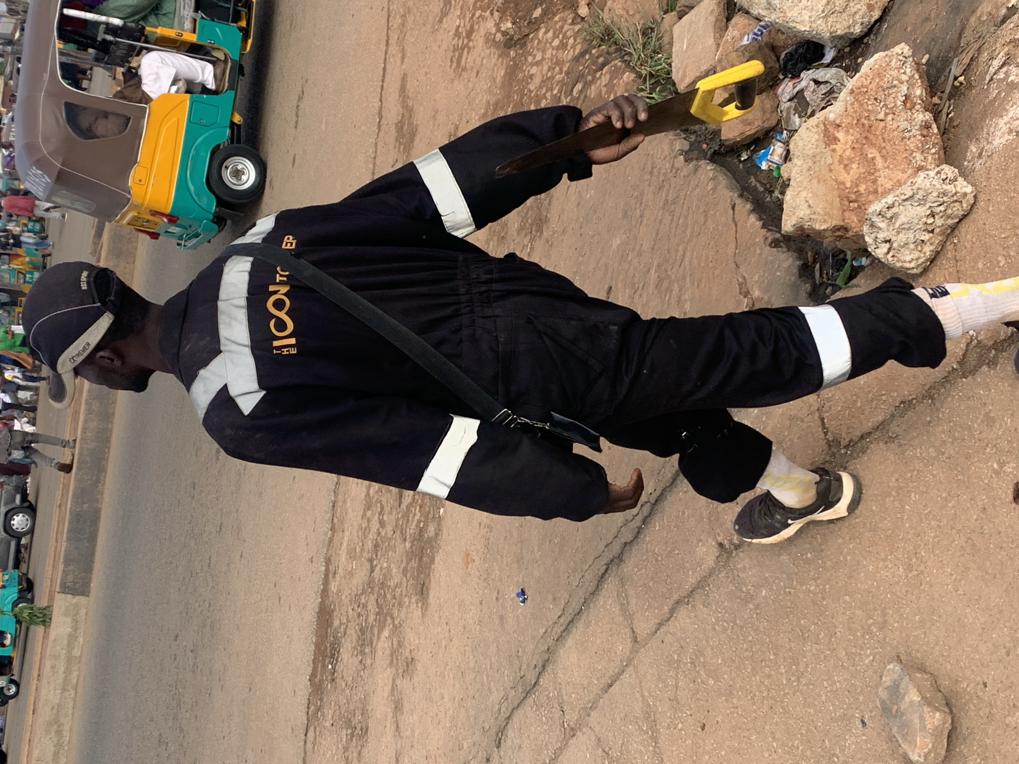 A person walks on a dirt and asphalt road in Gura Top, Nigeria, carrying a saw. The person is wearing a black jumpsuit with reflective silver stripes on the sleeves and chest. They also wear a black baseball cap and white socks with yellow accents visible above black sneakers. The person is walking away from the camera, and to their right, there is a pile of rocks and trash. In the background, a colorful auto-rickshaw is visible on the road, along with other people and vehicles. The environment appears to be an urban setting with evidence of commercial activity and traffic.