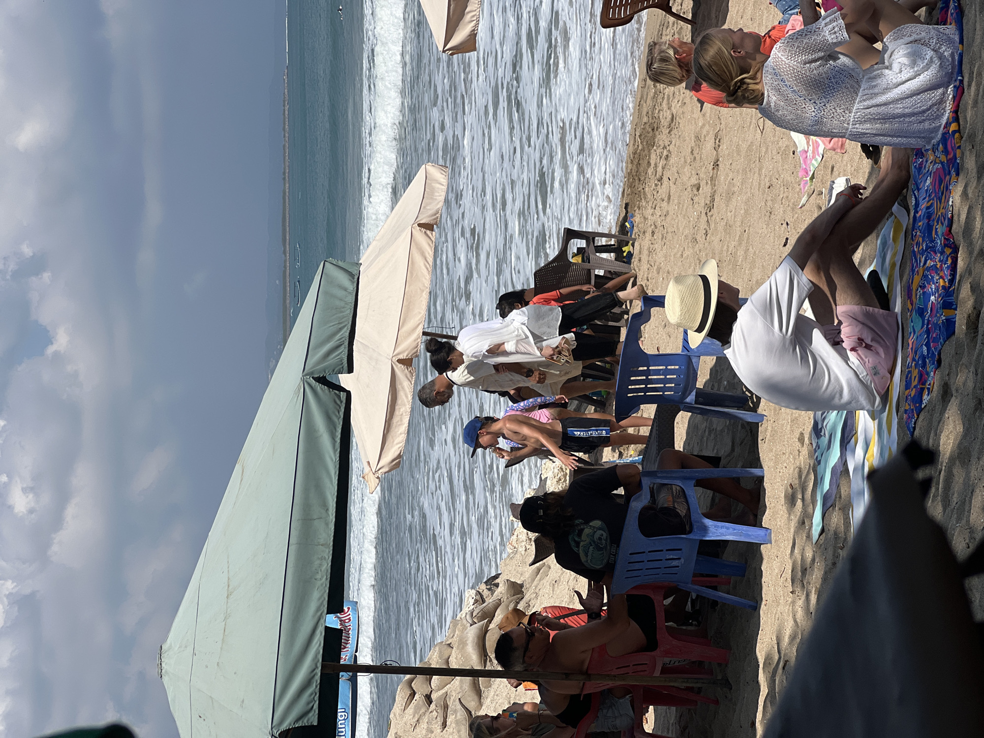In Kuta, Indonesia, a beach scene unfolds under a partly cloudy sky. Several people are gathered on the sand, seeking shade under large umbrellas. One umbrella, a light beige, is positioned over a group of individuals. Another, a dark green umbrella, casts a shadow over a sandy embankment reinforced with sandbags.
The ocean, with moderate waves, stretches to the horizon where a faint outline of land is visible. A few small boats are scattered on the water.
On the beach, a boy wearing a blue cap and swim trunks is seen. Nearby, an adult man in a black t-shirt with a graphic print is seated. Further back, a woman in a white top and black pants walks, carrying a pair of shoes. In the foreground, a man wearing a straw hat and light-colored shorts sits on a colorful towel, facing away from the camera. To his right, a woman in a white lace dress is seated on a towel.
Chairs of various colors, including red and blue, are placed on the sand. The overall atmosphere suggests a leisurely day at the beach.