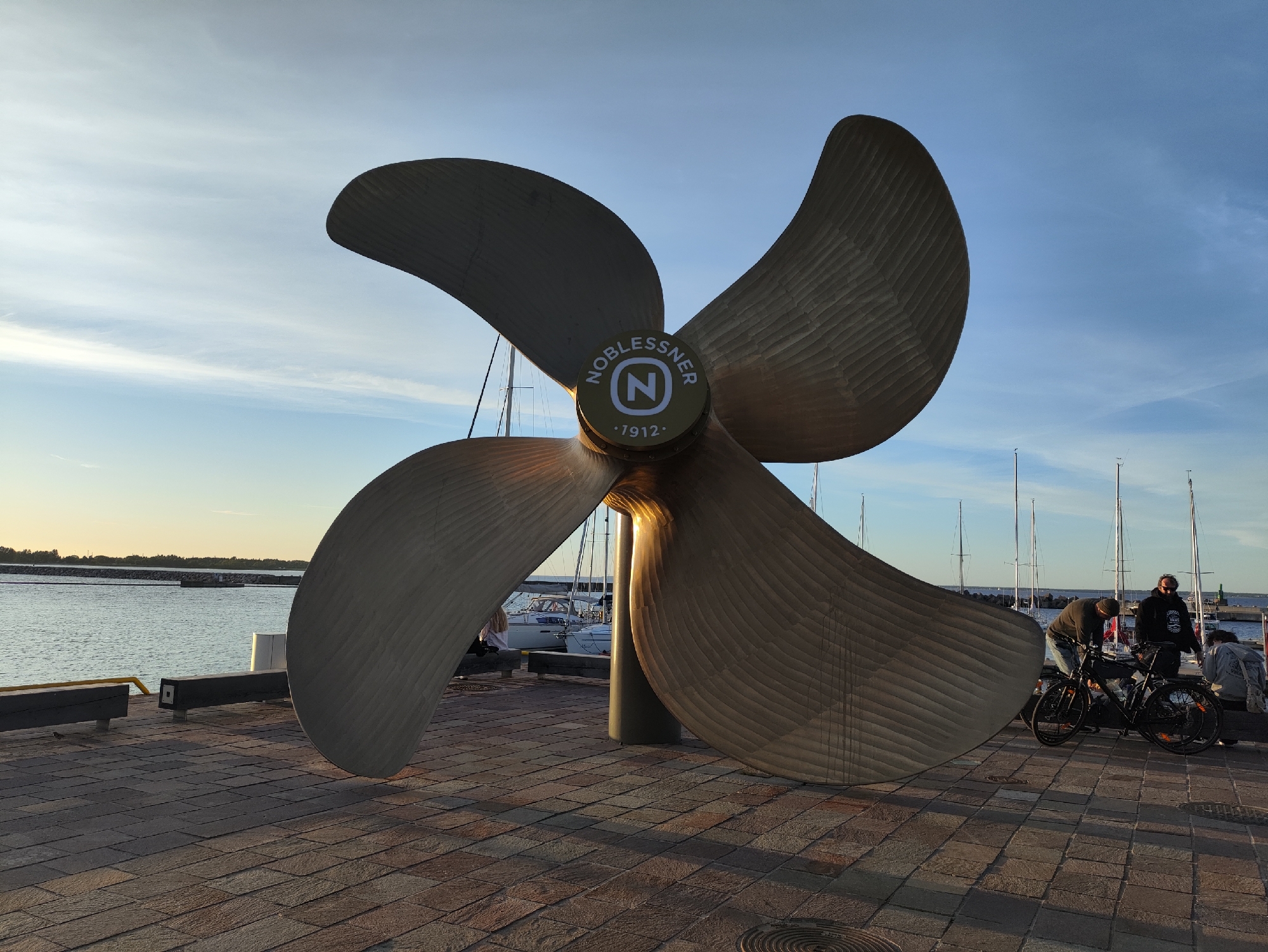 The image depicts a large metal propeller sculpture located on the waterfront promenade in the Noblessner district of Tallinn, Estonia. The propeller is made of a bronzed metal and has four blades. In the center of the propeller, there is a circular emblem with the inscription "NOBLESSNER" and the year "1912" surrounding a stylized letter "N".
In the background, several sailboats are moored in a harbor, and the calm sea extends to the horizon. The promenade is paved with rectangular stone tiles. Two dark wooden benches are visible on the left side of the image.
In the mid-ground, near the propeller, there are three people. One individual is bent over a black bicycle, appearing to be adjusting it. Another person stands nearby, looking at the bicycle. A third person is seated on a bench in the background, and another individual is partially visible in the far right. The sky is clear with a few wispy clouds, and the setting sun casts a warm glow on the scene.