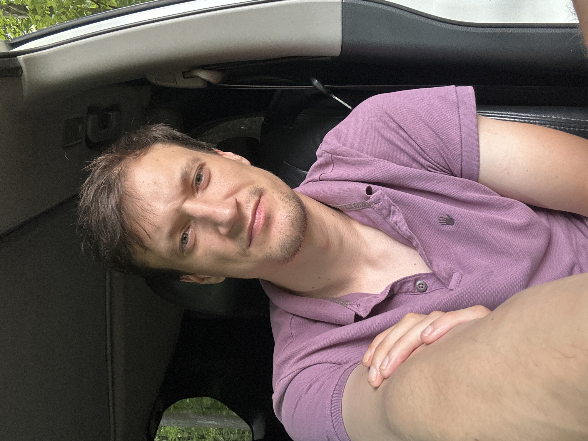 The image shows a close-up selfie of a man sitting in a car. He is wearing a purple polo shirt. The man has light skin, short brown hair, and a light stubble. He is looking directly at the camera with a neutral expression. His left arm is bent, with his hand resting on his bicep. The interior of the car is visible behind him, including a black leather seat and the car's headliner. Sunlight is filtering in from the side, suggesting it is daytime. The background outside the car window shows greenery, indicating a natural or rural setting. The exact location in Shatava, Ukraine, is not discernible from the image alone.