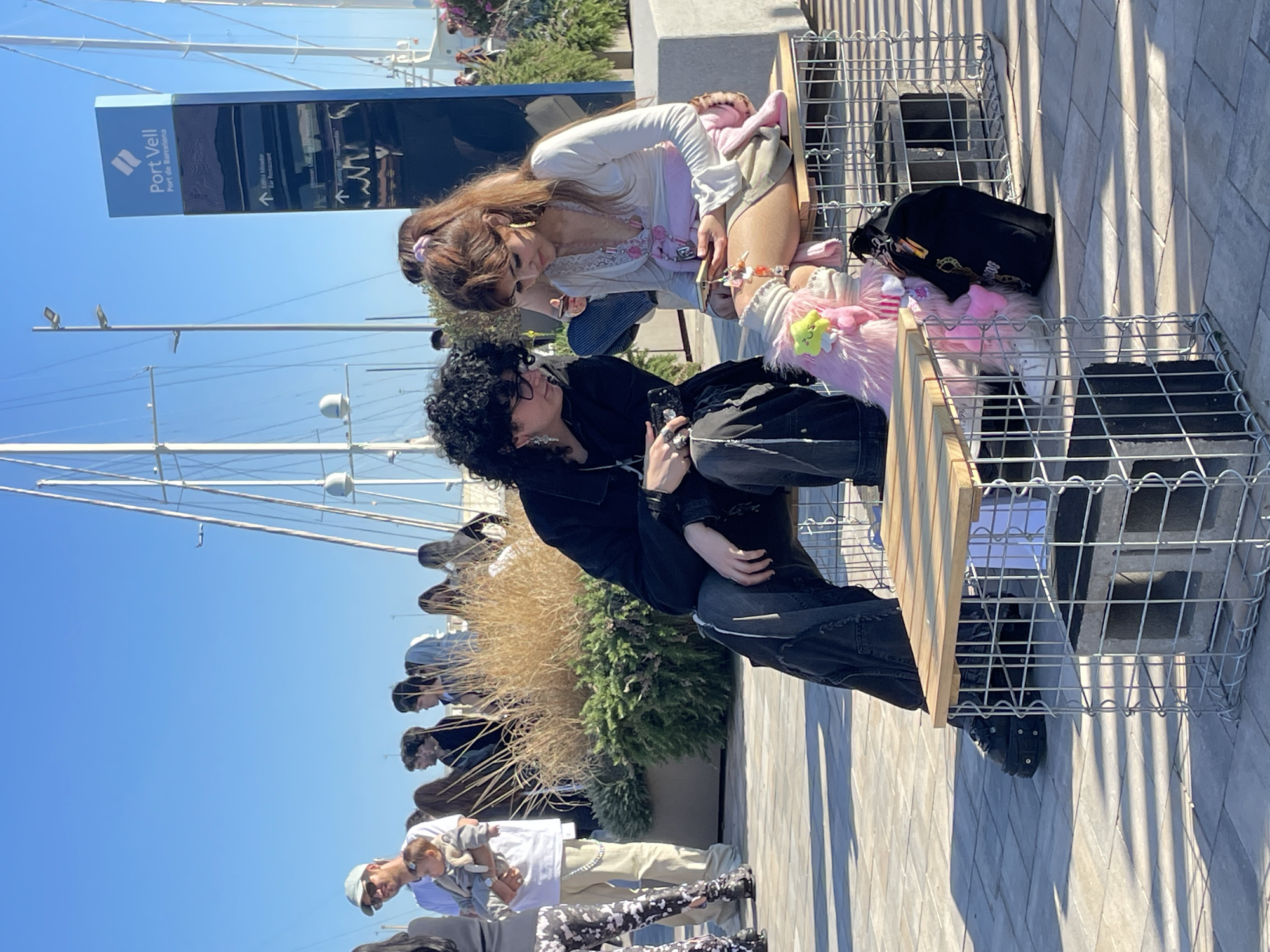 The image captures an outdoor scene on a bright, sunny day, likely a promenade or port area in Barcelona, Spain, given the visible "Port Vell" sign.
In the foreground, two individuals are seated on a gabion-style bench, which consists of a wire cage filled with rocks, topped with wooden planks. A woman with long brown hair, wearing a white long-sleeved top with lace details and light-colored shorts, is leaning forward, looking down at a phone she holds. Next to her, a man with curly dark hair, wearing a black jacket and dark pants, is also seated, holding a phone. They appear to be engaged with their devices or in a casual conversation. A black bag with a rainbow-colored strap and a fluffy pink accessory are placed near the woman on the bench.
The background features a clear blue sky. On the left, several boat masts are visible, indicating a nearby marina or harbor. A tall signpost stands prominently, displaying "Port Vell" and "Port de Barcelona" in white text on a blue background. Below this, smaller text reads "Edifici Mimamar" and "Bar Restaurant" with directional arrows. Other people are visible further in the background, some walking, including one person holding a baby. Architectural details include paved ground with strong shadows cast by structures and people, suggesting midday sunlight. Green and dried bushes are also seen in planters.