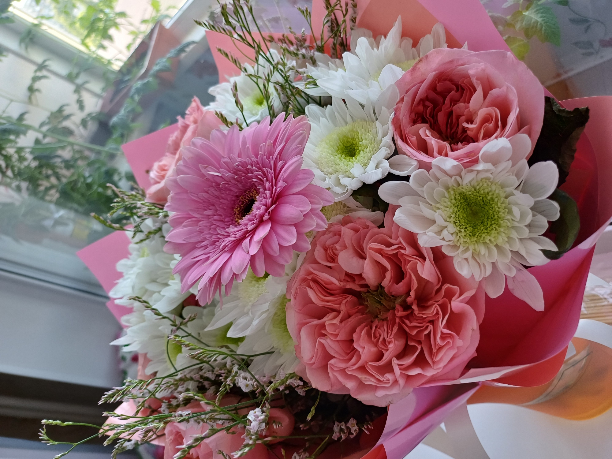 A close-up shot captures a vibrant bouquet of flowers, predominantly featuring soft pinks and whites, set against a backdrop of natural light filtering through a window. In the foreground, a large, intricately ruffled pink rose takes center stage, its petals layered and delicate. To its right, a bright pink Gerbera daisy with a dark center adds a pop of vivid color. Interspersed among the roses and Gerbera are clusters of white chrysanthemums with green centers, their petals radiating outwards. Delicate sprigs of greenery and small white wildflowers add texture and depth to the arrangement. The bouquet is partially wrapped in layers of pink and orange paper, suggesting it is a gift. The background is softly blurred, hinting at a window with greenery visible outside, implying an indoor setting near a natural light source. The lighting is bright and diffused, highlighting the textures and colors of the flowers.