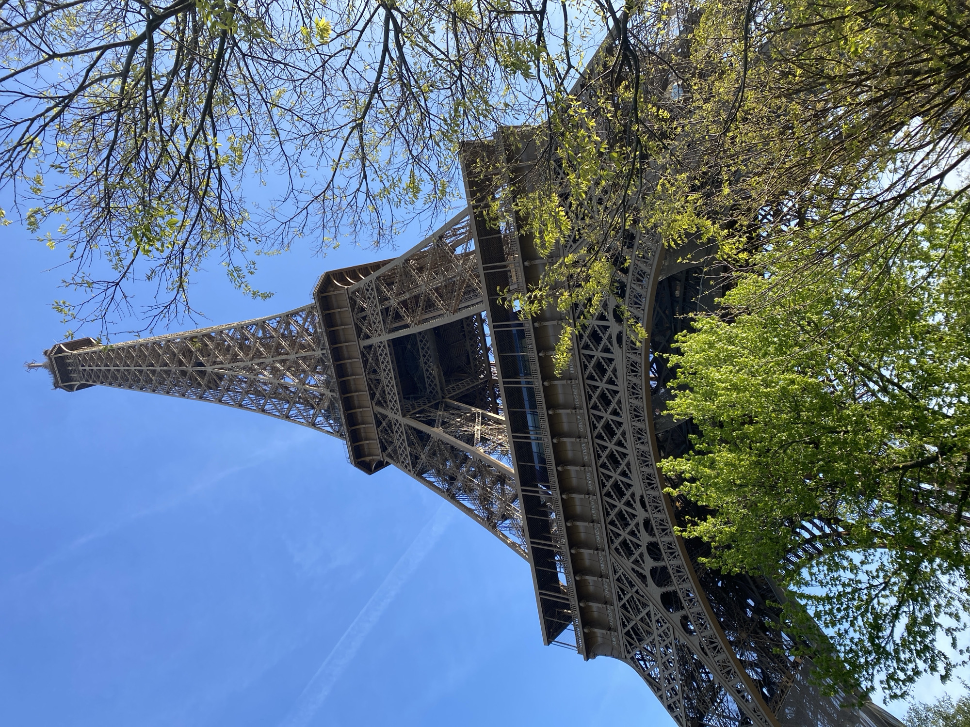 The Eiffel Tower, an iron lattice tower on the Champ de Mars in Paris, France, is shown from a low angle. The tower is visible against a blue sky with wispy clouds. Trees with green leaves partially frame the tower on both sides. The tower appears to be constructed of an open lattice work of metal beams. The angle is angled, giving the impression the tower is very tall.