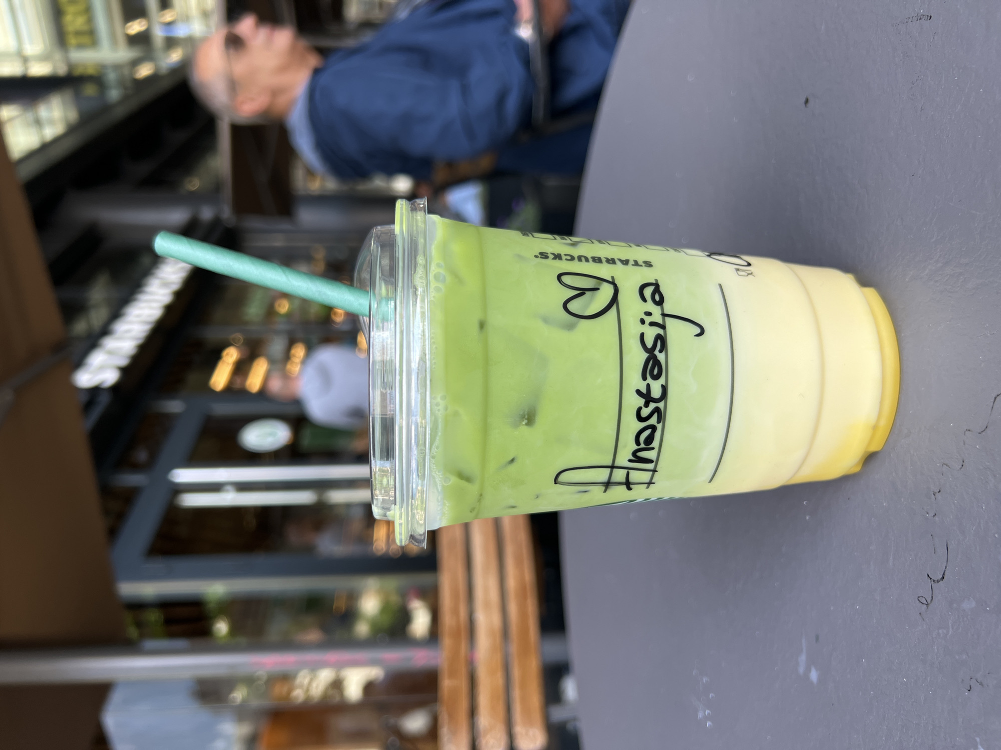 Here's a description of the image:
**Subject:** The primary subject is a Starbucks iced matcha latte with a blue-green paper straw. The cup has the name "Anastazija" written on it with a heart symbol. The drink appears to have two layers, the top being a green matcha mixture and the bottom being a creamy yellow liquid.
**People:** A blurry person is standing in the background, and another is behind glass, closer to the drink.
**Location:** The drink is on an outdoor table in front of a Starbucks store in Belgrade, Serbia.
**Environment:** The image is taken outside, as indicated by the natural lighting. The background shows a building with glass windows and doors, which is likely the Starbucks store front. There are wooden chairs around the table. The weather appears to be mild.
**Objects:**
* Starbucks cup with matcha latte
* Paper straw
* Table
* Chairs
* Starbucks storefront
**Actions:** The action is the presence of the drink on the table and the presence of other people in the background.