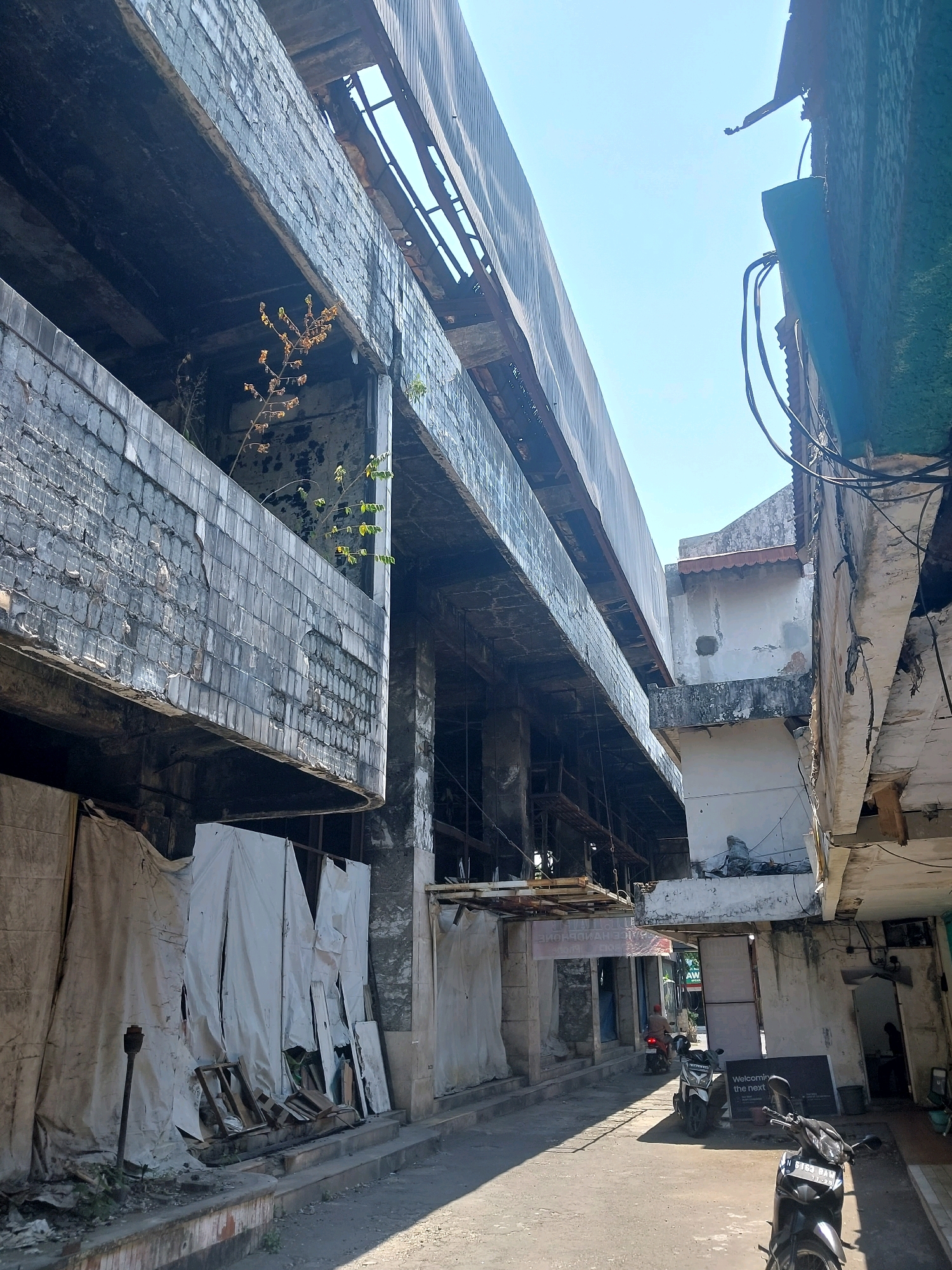 The image depicts a narrow street in Malang, Indonesia, flanked by an old, partially dilapidated building on the left and other structures on the right. The building on the left appears to be an abandoned or under-renovation structure, with exposed concrete, broken tiles, and scaffolding visible. White tarps are draped over parts of the lower levels, suggesting ongoing work or an attempt to shield the interior. A small tree grows from an upper level of the building.
On the right side of the street, a two-story building shows signs of age, with peeling paint and exposed wiring. In the mid-ground, two scooters are parked on the street, one in front of a sign that reads "Welcoming the next." Further down the street, a person is seen riding a scooter. The street itself is paved and shows areas of sunlight and shadow, indicating a bright, sunny day. The overall scene conveys a sense of urban decay and a busy, yet somewhat neglected, streetscape.