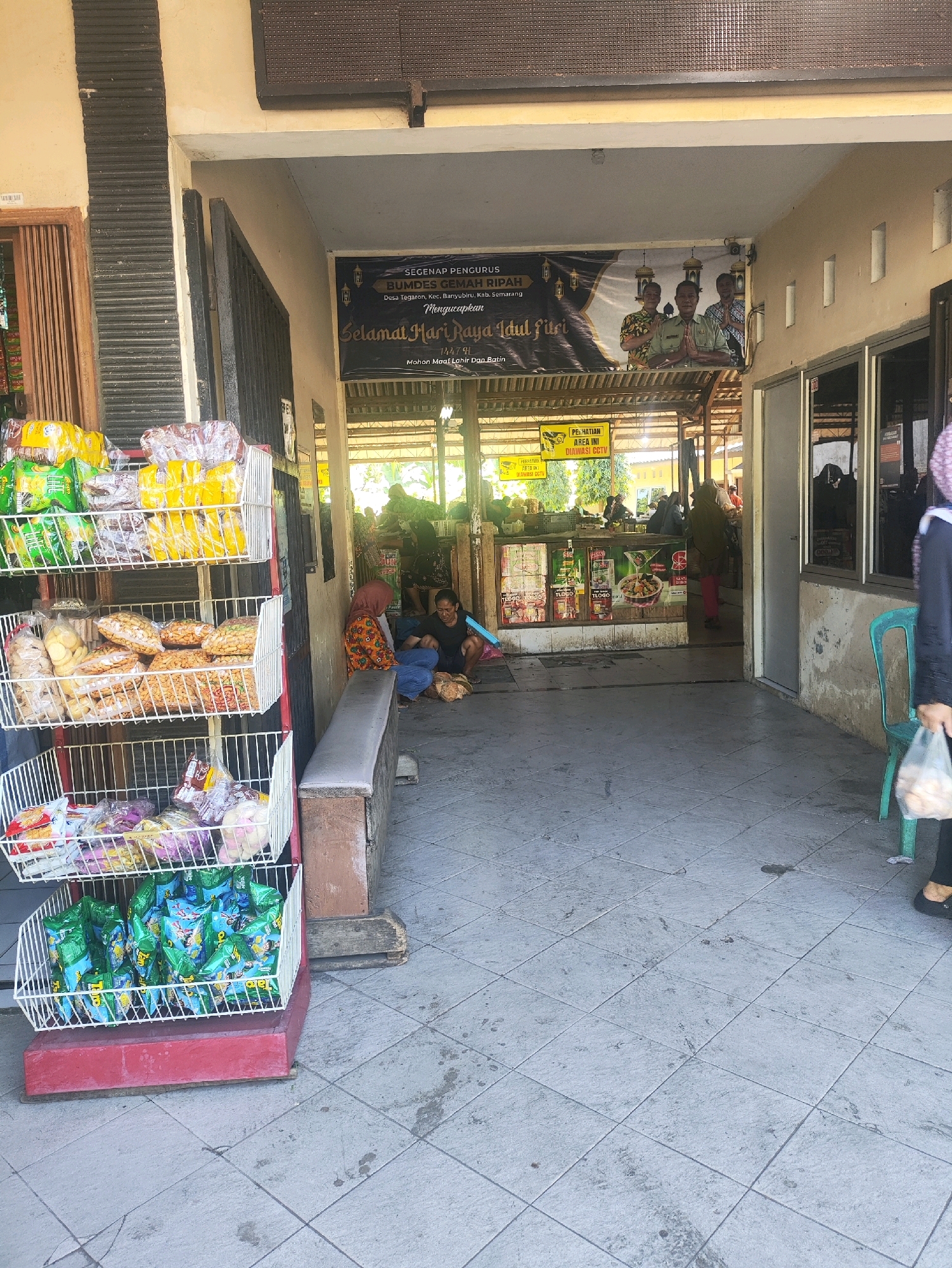 This image depicts a street-level view of a shop entrance in Krajan Tegaron, Indonesia. In the foreground, a multi-tiered white wire rack displays various packaged food items, including snacks and candies. To the left of the rack, a wooden and metal structure is visible. Adjacent to this is a covered walkway leading into a shop or food stall. Inside the shop, several people are present, some seated and others standing. A banner hangs prominently above the entrance, bearing text in Indonesian, including "Selamat Hari Raya Idul Fitri" (Happy Eid al-Fitr). The shop interior appears to be stocked with various goods. The floor is paved with square tiles, some of which are marked with dirt or stains. A green plastic chair is positioned outside the shop on the right. The overall environment suggests a commercial area, likely a small local business.