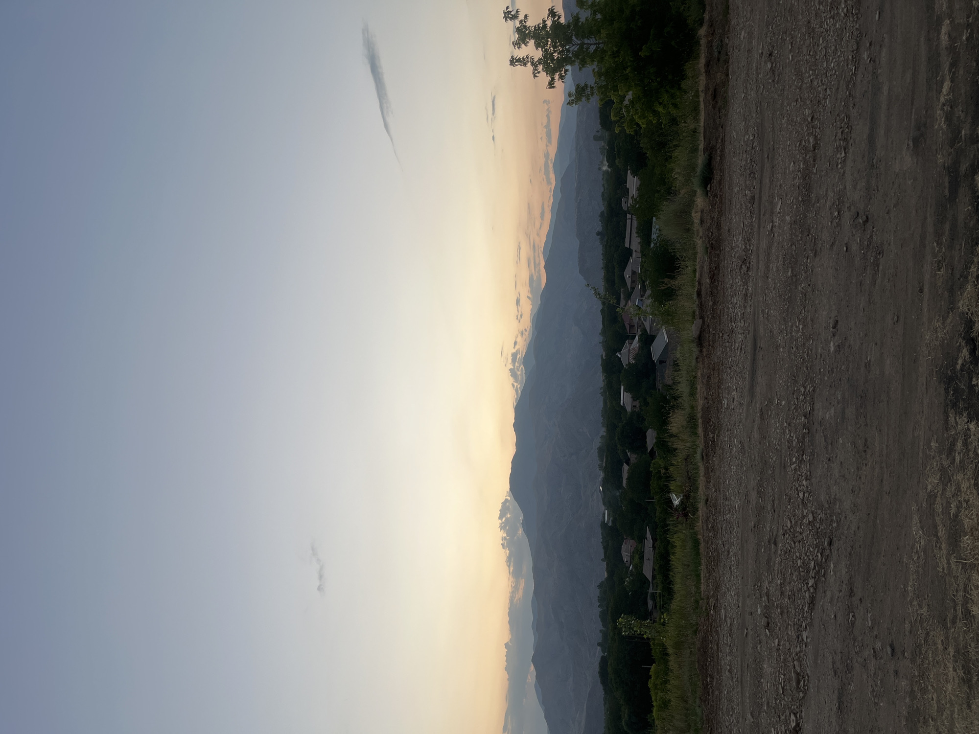 A wide-angle, natural light photograph captures a serene rural landscape at dusk, likely in or near Yeghegnadzor, Armenia. The foreground is dominated by a rough, dirt road or path, textured with loose gravel and dry grass. This path leads uphill towards a more cultivated area.
In the midground, a scattering of houses with dark roofs are nestled amongst lush, green trees. The village appears quiet and residential. Beyond the village, a range of majestic, layered mountains stretches across the horizon, their forms softened by the twilight.
The sky above is a gradient of soft blues and pale oranges, indicating the sun has recently set, with a few wispy clouds scattered across the expanse. The overall atmosphere is one of peacefulness and natural beauty, with no people or animals visible. The scene is captured from an elevated position, looking down upon the village and mountains.