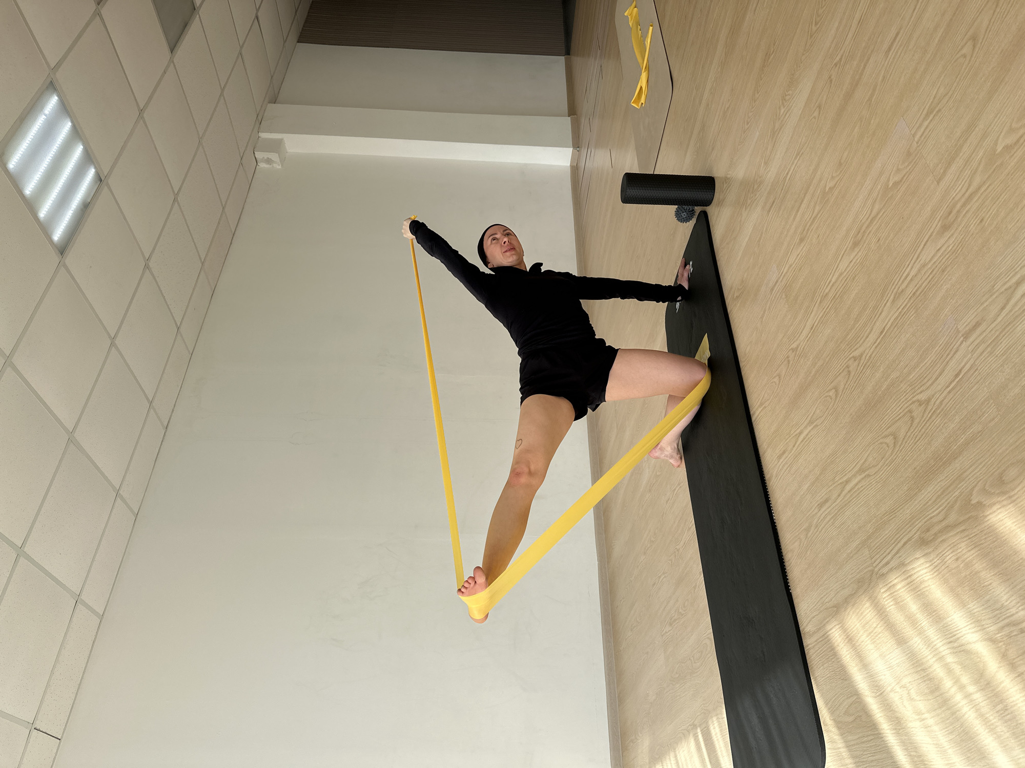 A female individual is performing an exercise with a yellow resistance band in an indoor setting, likely a gym or studio, in Votkinsk, Russia. The image is taken from a low angle, looking up at the person, who is centered in the frame. She is dressed in a black long-sleeved top, black shorts, and a black beanie.
The woman is engaged in a resistance band exercise. Her left foot (from the viewer's perspective) is planted on a black yoga mat, which lies on a light-colored wooden floor. Her right leg is extended outwards, with one end of the yellow resistance band looped around her bare foot. The other end of the band is held in her right hand, which is extended upwards and slightly to her side, creating tension in the band. Her left arm is slightly bent, with her hand resting on her hip or upper thigh. She appears focused, looking straight ahead or slightly upwards. A small, heart-shaped tattoo is visible on her left thigh.
On the wooden floor to her right, beside the yoga mat, are a black foam roller and a small black spiky massage ball. The room features plain white walls and a suspended ceiling with white square acoustic tiles. One section of the ceiling includes a brightly lit fluorescent light fixture. Diagonal shadows are cast across the wooden floor on the right side of the image, suggesting natural light from an unseen window, indicating it is daytime. No visible text is present in the image.