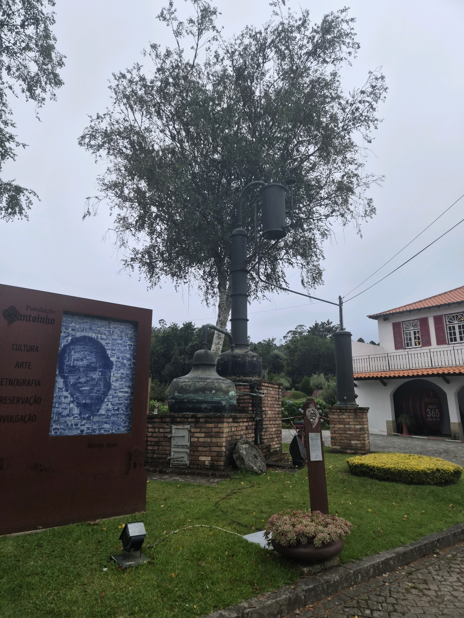 The image shows a historical artifact displayed outdoors, likely in a park or museum setting. In the foreground, a tall, weathered metal structure resembling a still or distillation apparatus stands prominently. It is surrounded by a low brick wall, with a large, dark-colored sign to the left. This sign features text and a blue and white mosaic portrait.
The sign reads "Fundação Santoinho" at the top, followed by "CULTURA," "ARTE," "ETNOGRAFIA," "RESERVAÇÃO," and "DIVULGAÇÃO." The mosaic portrait appears to be of a man's face.
Behind the artifact and sign, there is lush green grass and various trees, suggesting an outdoor environment. To the right of the main artifact, another similar, though smaller, metal structure is visible. Further to the right, a white building with red-shuttered windows and a terracotta tiled roof is situated. A balcony with a railing overlooks an arched entrance where a large barrel-shaped sign reads "Santoinho Aberto 365 Dias No Ano" (Santoinho Open 365 Days a Year).
The sky is overcast and pale, indicating a cloudy day. The overall impression is of a historical site or cultural establishment dedicated to preserving and presenting local heritage. The presence of the "Fundação Santoinho" sign strongly suggests this is located at the Santoinho Foundation in Darque, Portugal.