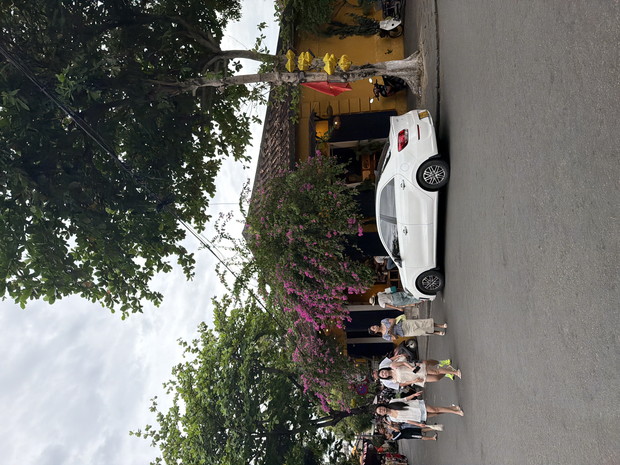 The image depicts a street scene in Hoi An, Vietnam, under an overcast sky. A white sedan is parked on the right side of the asphalt road. Several individuals are walking on the sidewalk and street on the left. A building with a yellow facade and a tiled roof is visible behind them, adorned with vibrant pink bougainvillea. Lush green trees with dark leaves overhang the street. The scene conveys a sense of everyday urban life in a Southeast Asian city.