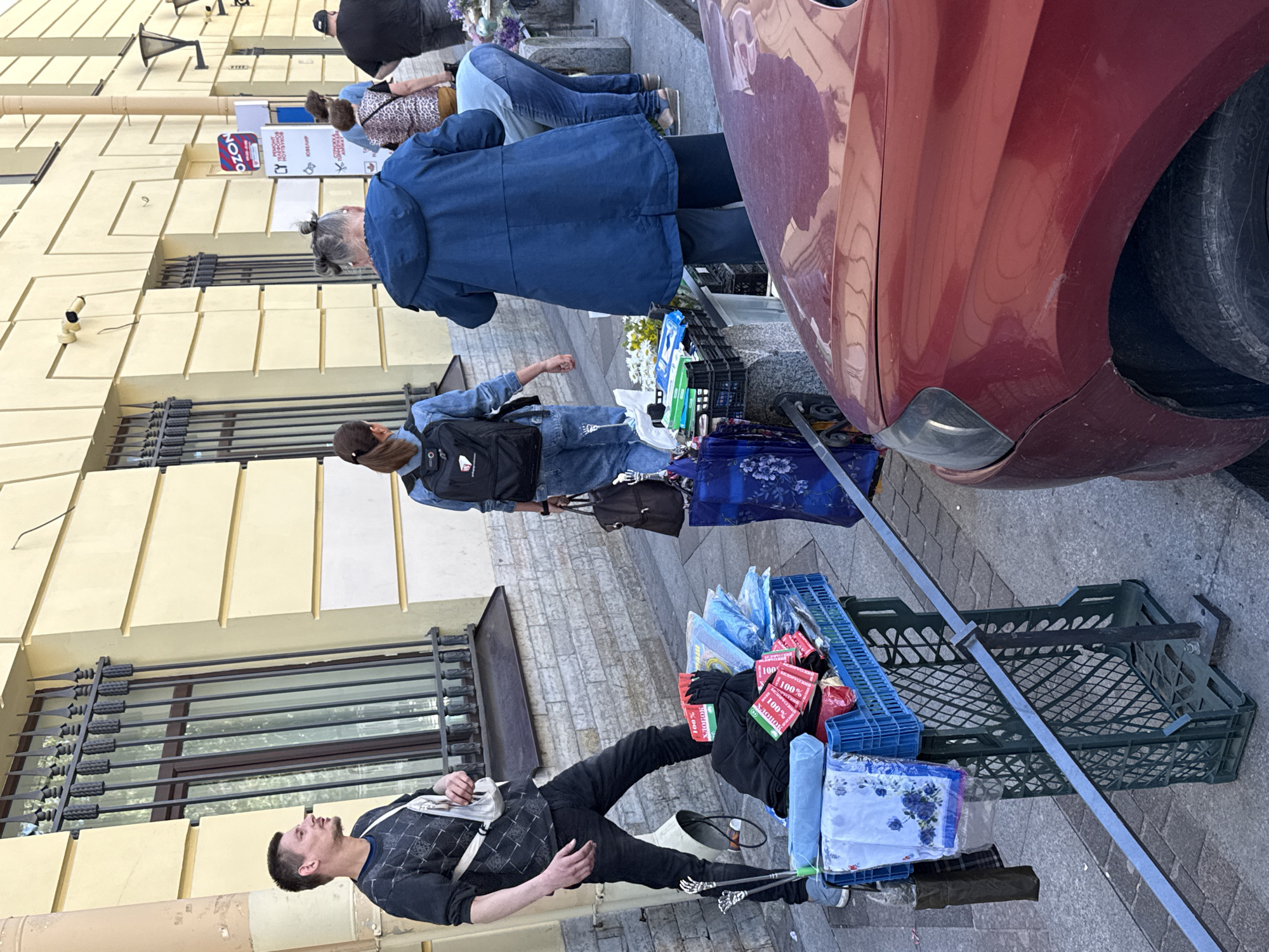 The image shows an outdoor street scene with several people and various objects. The primary subjects are individuals standing and walking on a sidewalk in front of a building. One person in the foreground is lying down, propped up by objects around them.
Various objects are visible, including shopping bags, plastic crates filled with items, umbrellas, and what appear to be fabric items or cloths. A red car is parked on the right side of the frame. The building in the background has a light yellow facade with dark window bars.
Text visible in the image includes "100%", "MOHOIX", "Хлопок", "OZON", and "СЮВЕЛИР". The presence of these signs, along with the collection of goods displayed in crates and bags, suggests a market or street vending activity.
The lighting indicates it is daytime, likely sunny given the clear shadows. The scene appears to be a casual street environment in a city, possibly in Russia given the Cyrillic text. The people's postures and the general ambiance do not suggest any particular strong emotions; it appears to be a typical day. The specific mention of Saint Petersburg provides further location context.