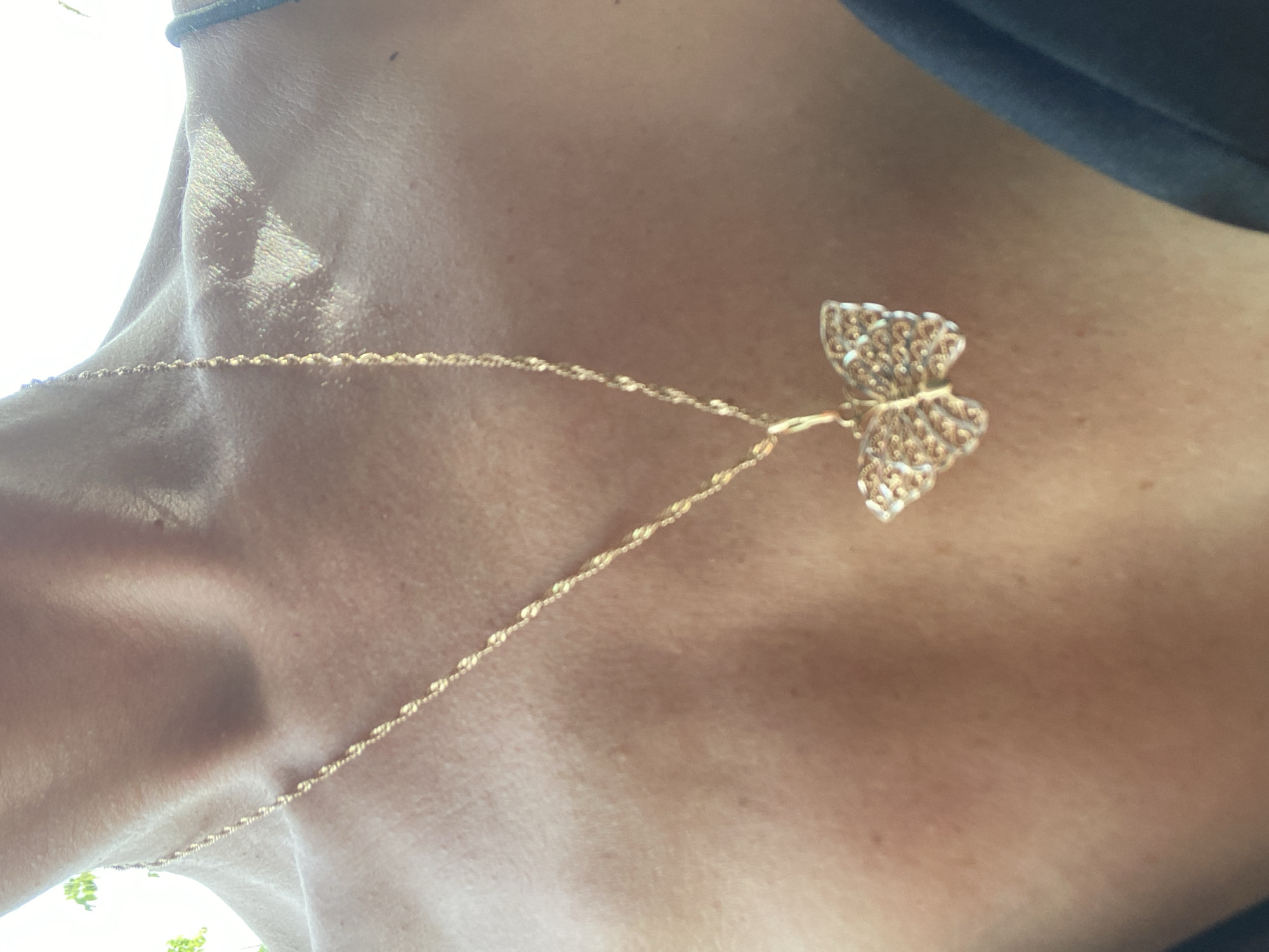The image shows a person's neck and upper chest wearing a gold-colored butterfly pendant necklace. The necklace is a thin chain with a butterfly-shaped pendant made of intricate metalwork. The person is wearing a dark-colored garment at the top of the image, suggesting they are indoors with bright light coming in from the side. The location is Baan Tai, Thailand.