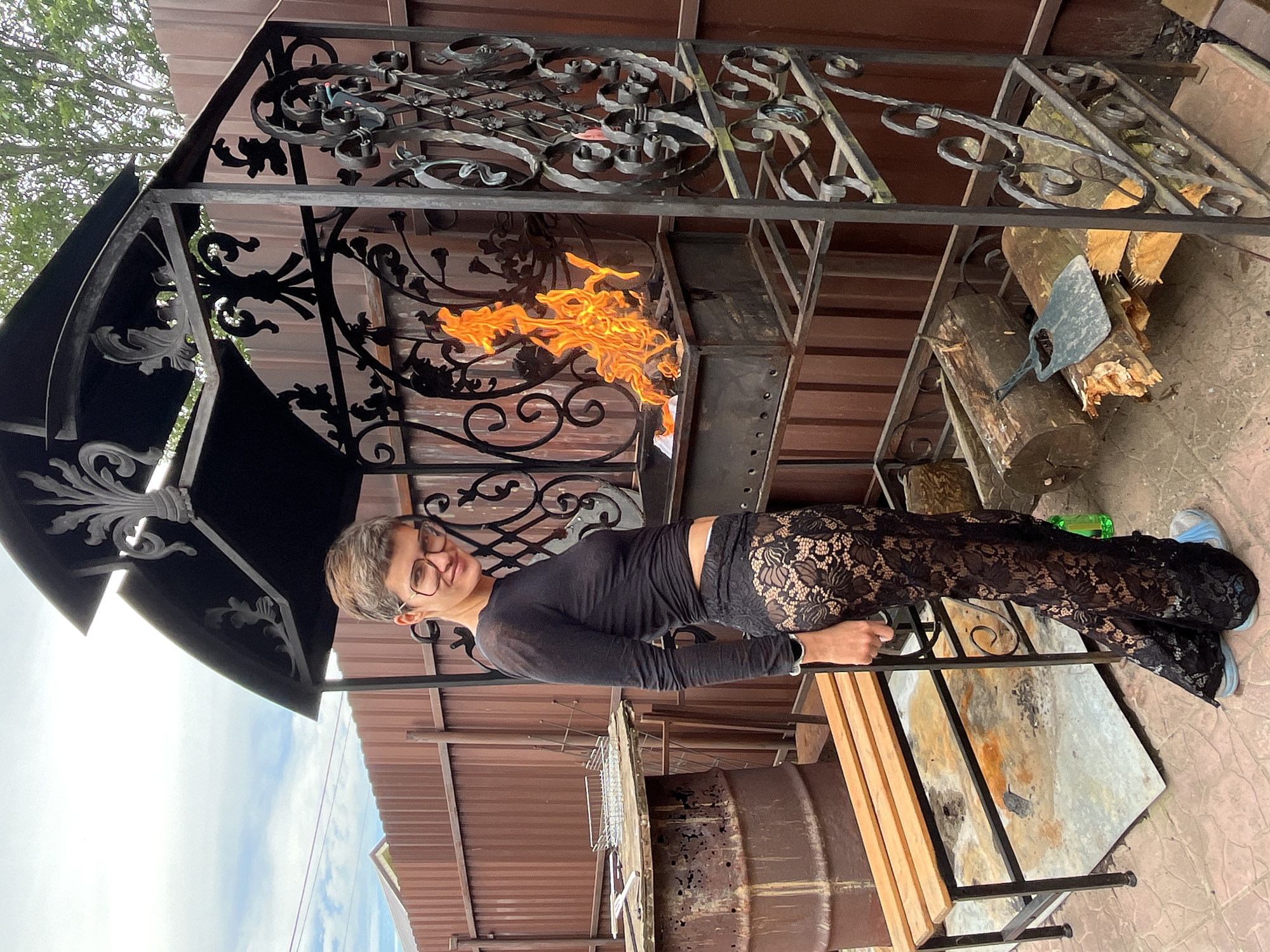 A woman with short, light brown hair and glasses stands next to a black metal barbecue in an outdoor setting. She is wearing a black long-sleeved shirt and black lace pants. The barbecue has a fire burning inside it, with flames visible. There are logs of wood placed on a lower rack of the barbecue, and a black tool with a scoop-like end rests on the logs. To the left of the barbecue, there is a wooden bench and a large, rusty metal barrel. In the background, a brown wooden fence and some greenery are visible. The ground appears to be paved with textured tiles. The scene is consistent with a backyard or outdoor cooking area in Вельяминово, Russia.