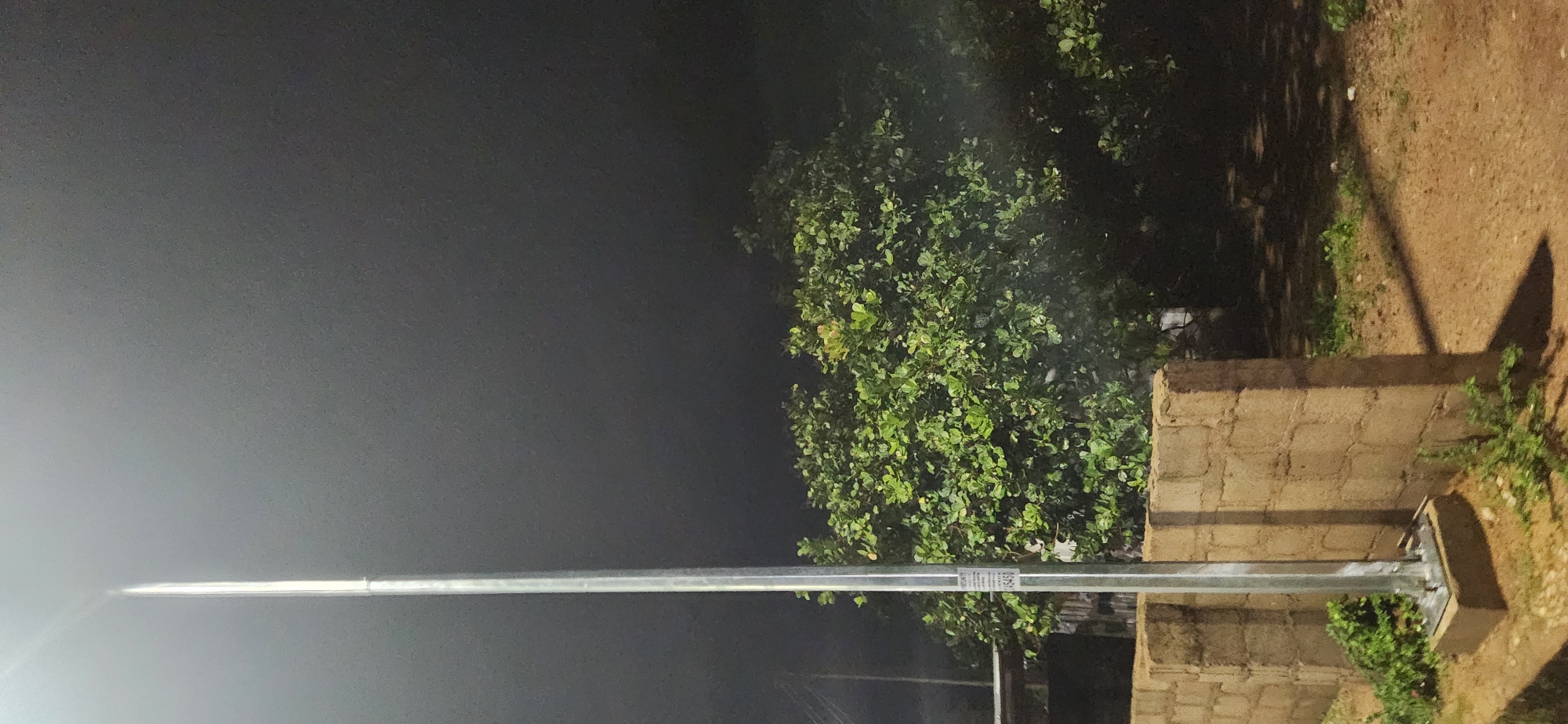 This is a nighttime, eye-level, medium shot of a metal lamppost standing next to a rough, cinderblock wall. The lamppost is to the left of the frame, and a large, leafy tree is positioned behind the wall. The sky is dark and overcast. The lamppost has a sticker affixed to it with text and numbers. The wall is constructed of rectangular cinder blocks and shows signs of wear and tear. There is some sparse green vegetation growing at the base of the wall and around the lamppost. The ground is covered in dirt and scattered pebbles. The image appears to have been taken in an outdoor setting.