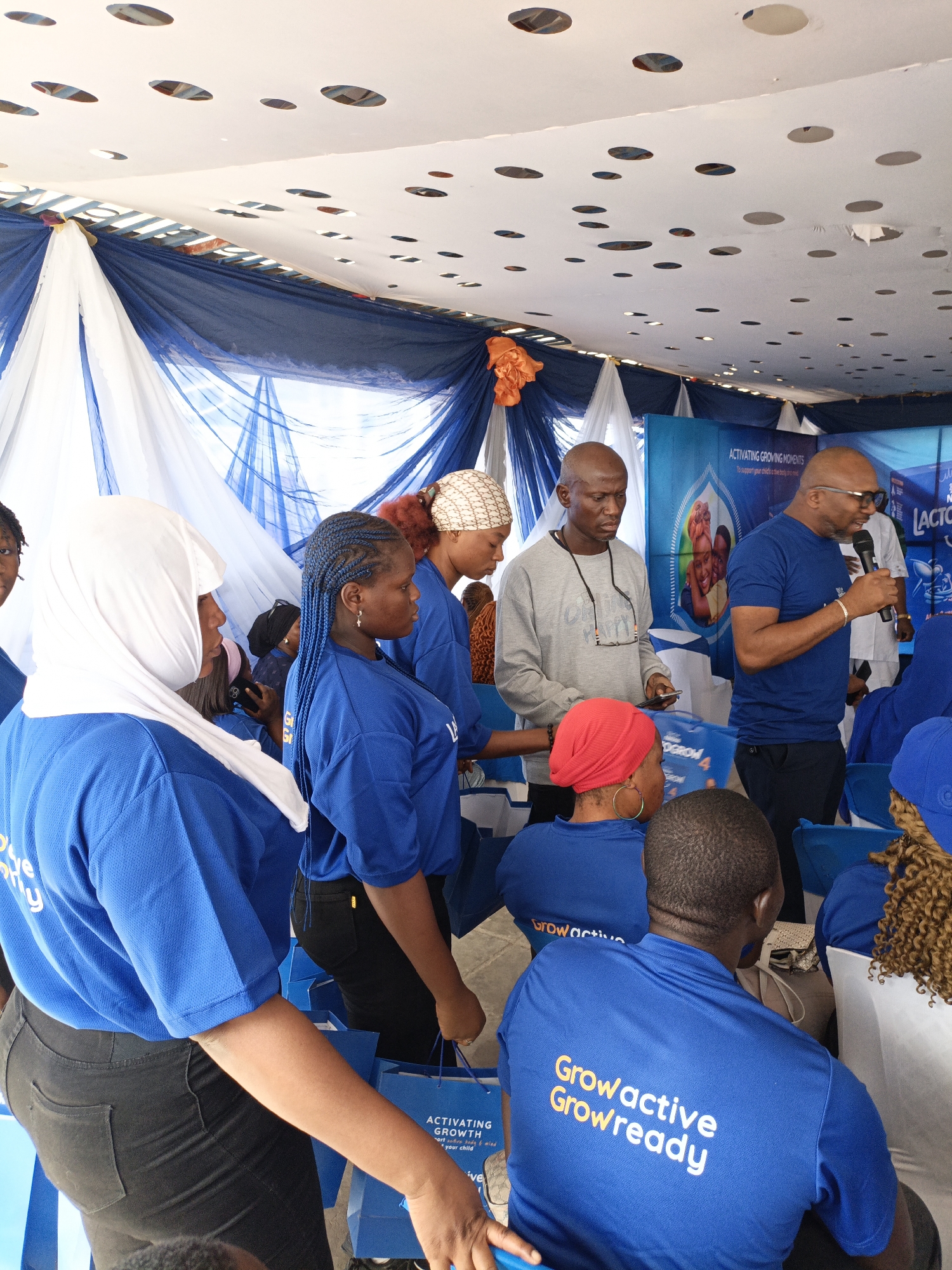 Here is a detailed description of the image:
**Scene:**
The image appears to be taken inside a tent or a room decorated for an event in Tarauni, Nigeria. The ceiling is covered with white and blue drapery, and there are circular cutouts in the material above.
**People:**
There is a group of people in the scene. Most are wearing blue shirts with "Growactive Growready" printed on the back, indicating they may be part of a promotion or event.
- At the front, a woman is wearing a white headscarf.
- Next to her is a woman with blue braided hair.
- Some people are sitting in chairs, listening to a speaker.
- Two men are standing towards the right, engaged in presenting or speaking. One is wearing a blue shirt and speaking into a microphone, while the other is wearing a gray shirt and holding a phone.
**Objects:**
- There is a banner in the background with the words "ACTIVATING GROWING MOMENTS" and an image of children.
- Bags with branding related to "Lacto Grow" are present.
**Actions & Interactions:**
- People are primarily engaged in listening and observing.
- One man is speaking into a microphone, addressing the audience.
- The other man is holding a phone and engaged in some activity, possibly related to the presentation.
**Environment:**
- The location is decorated, likely for a promotional or informational event.
- The presence of branding and promotional material suggests a marketing campaign or product launch.